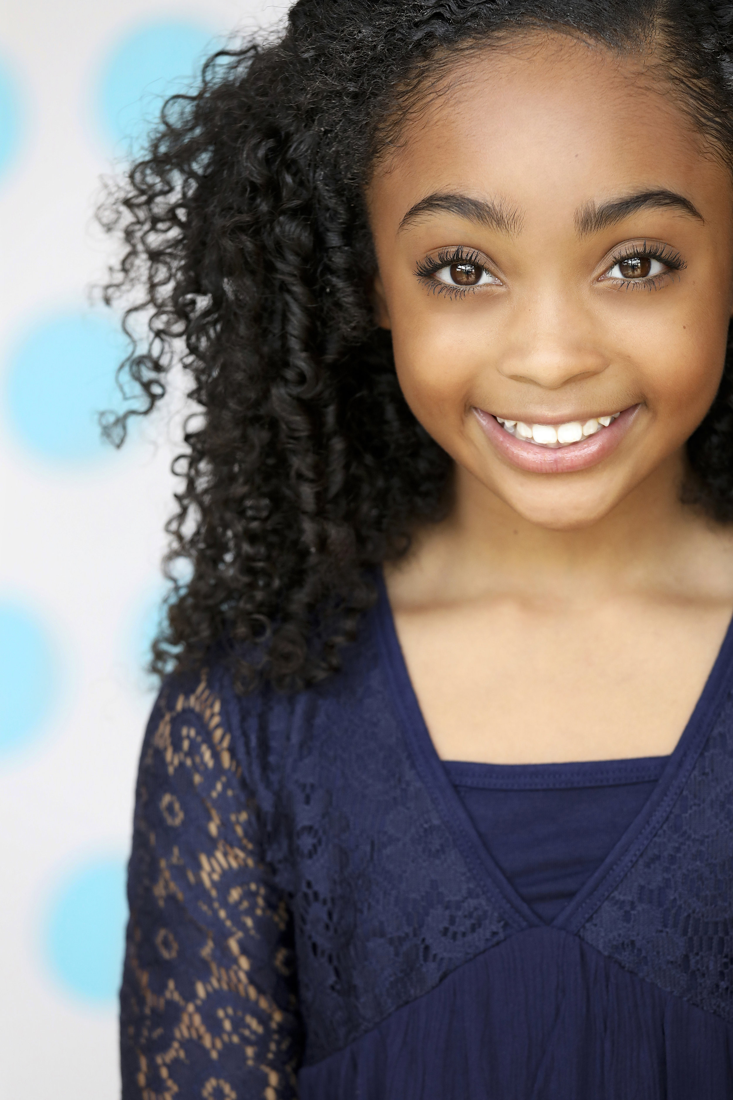 Close-up of a girl with curly black hair, brown eyes, and a big smile, wearing a navy blue lace top, standing in front of a light blue polka dot background.