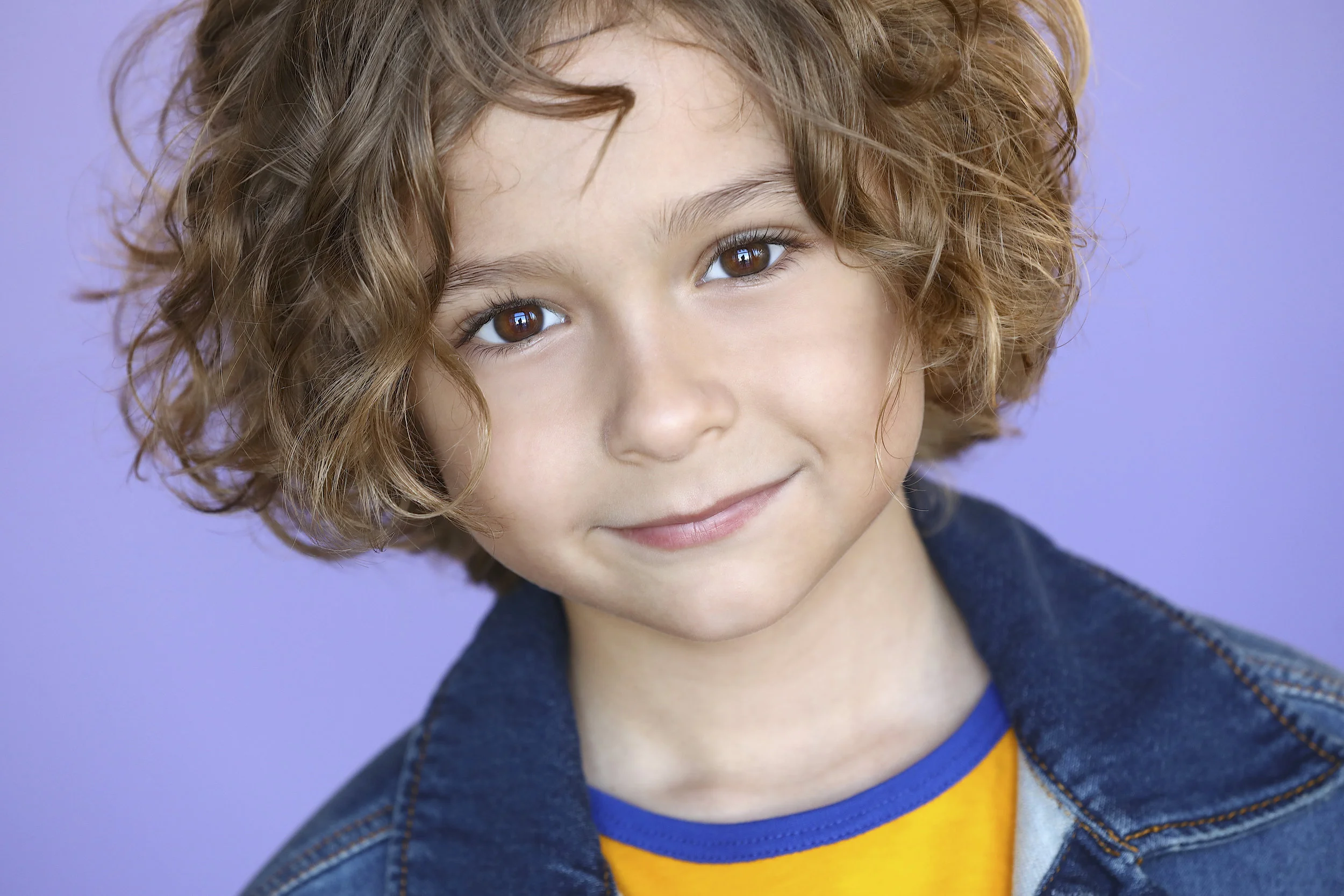 Close-up portrait of a young boy with curly, light brown hair and brown eyes, smiling gently, wearing a denim jacket and yellow shirt with blue trim, against a purple background.