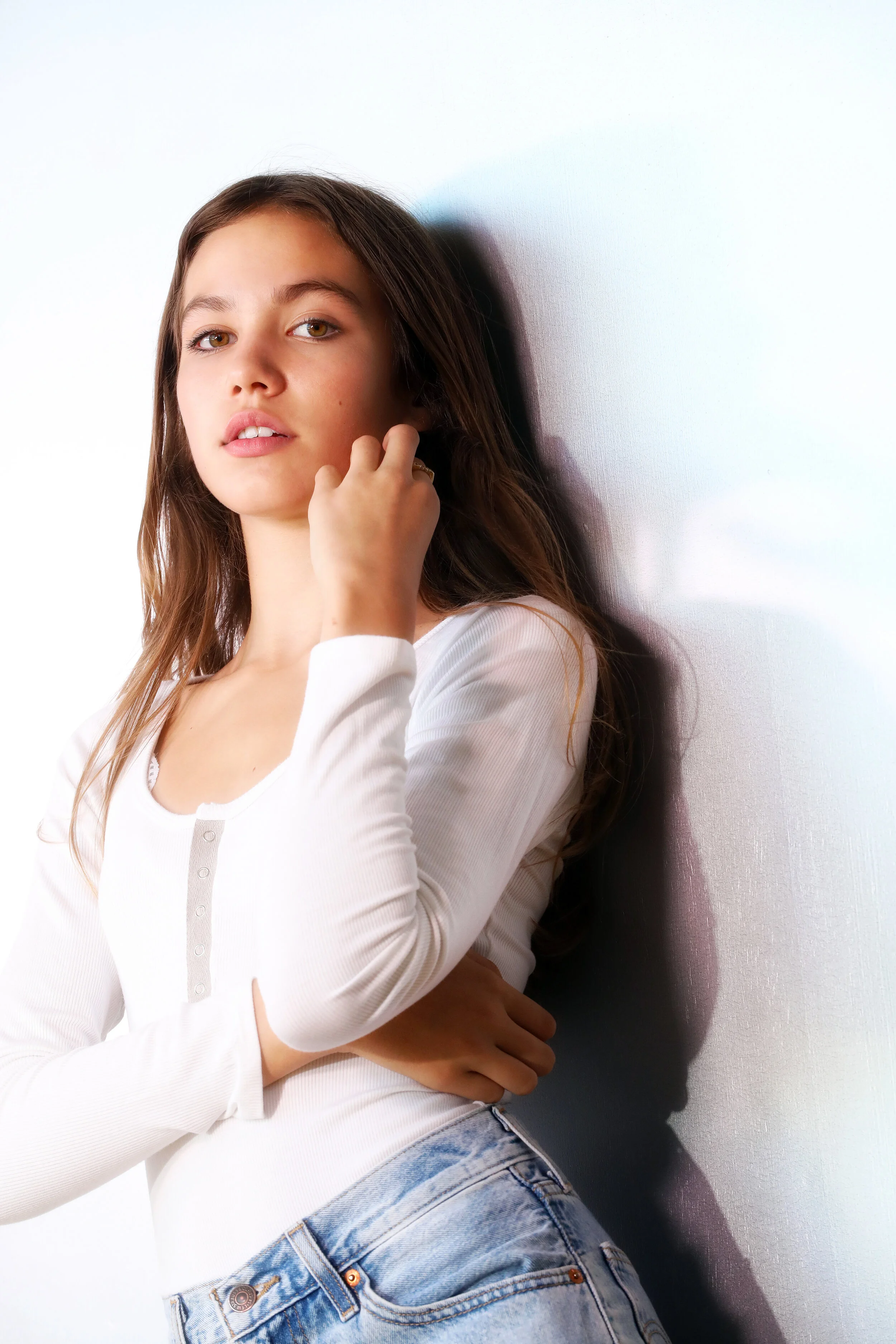 A young woman with long brown hair, wearing a white long-sleeve top and blue jeans, standing against a plain white wall.