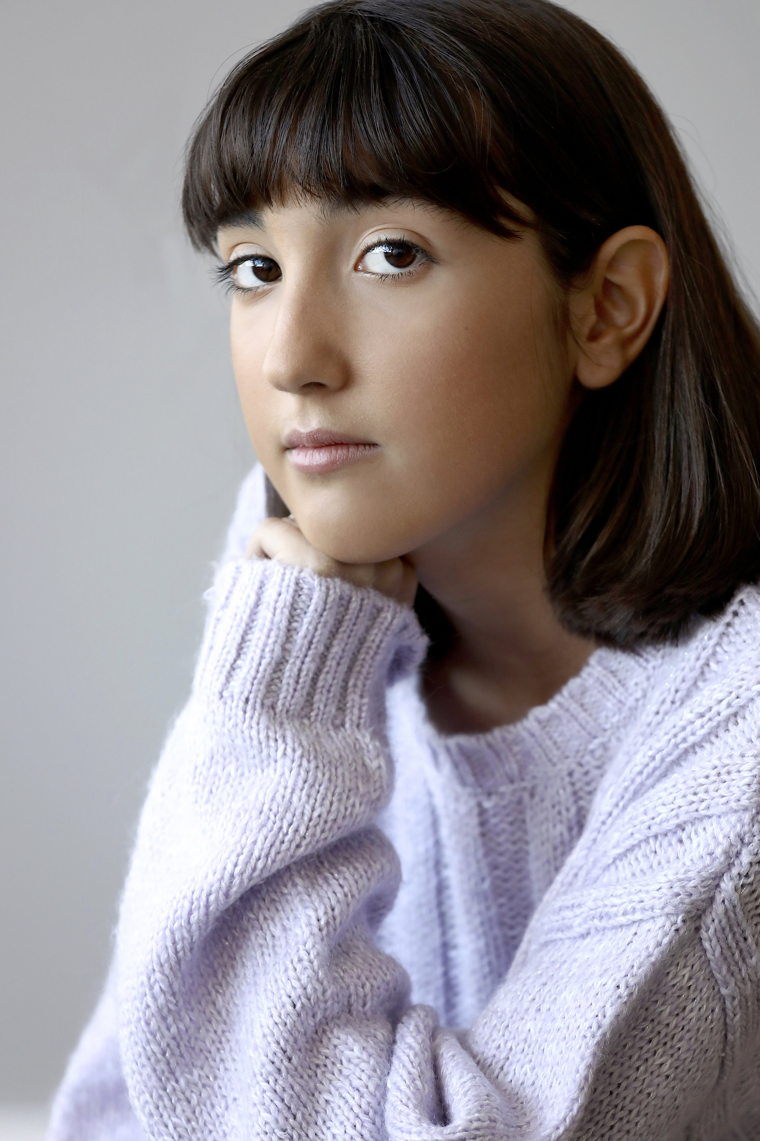 A young woman with brown hair and light skin looking at the camera, resting her chin on her hand, wearing a cozy light purple sweater.
