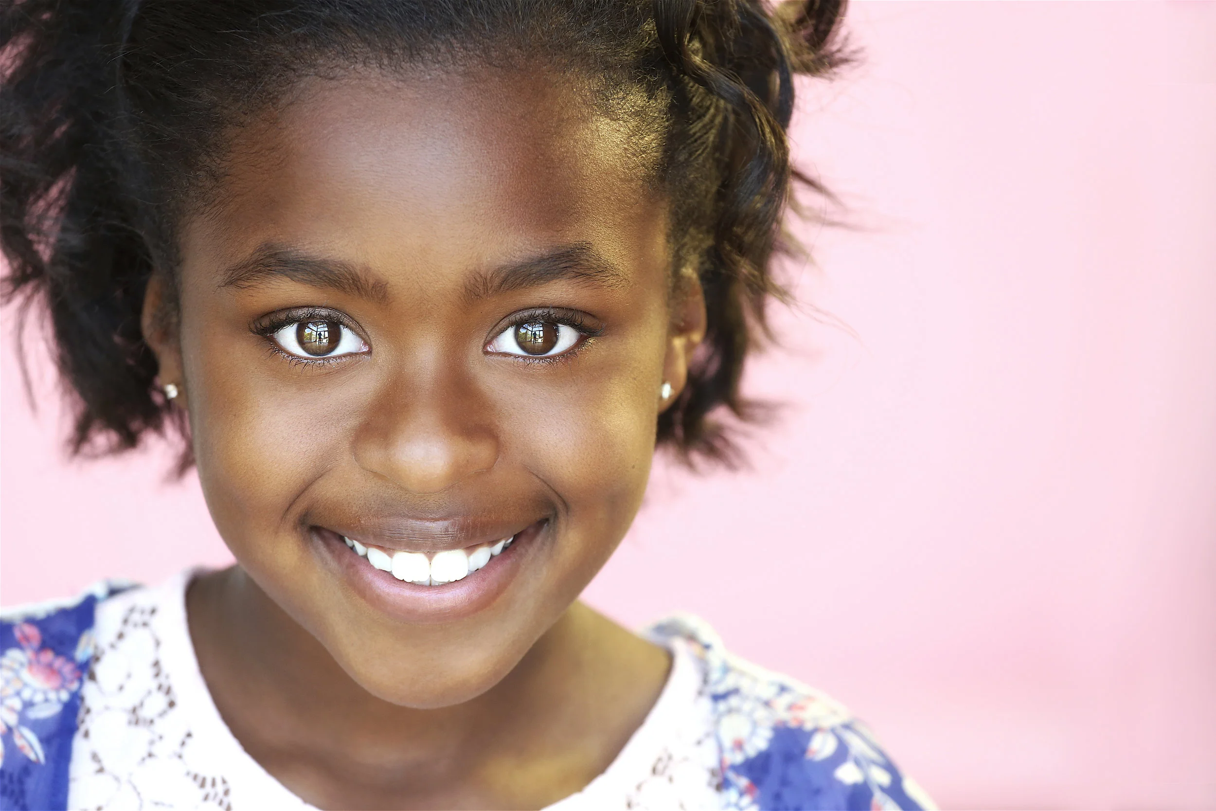 Close-up of a young girl with brown skin, large brown eyes, smiling, wearing pearl earrings and a blue floral dress against a pink background.