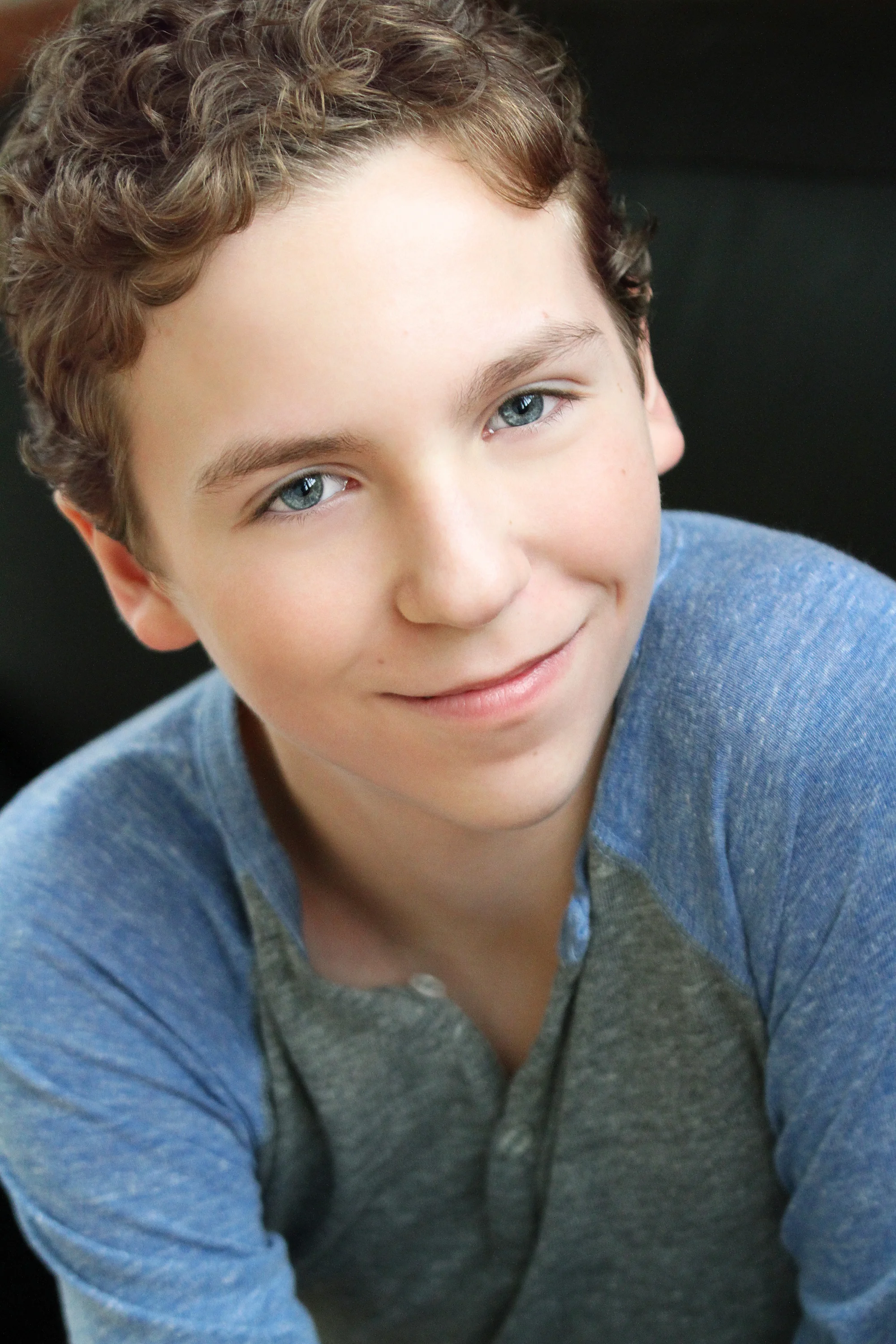 Close-up of a smiling teenage boy with curly brown hair and blue eyes, wearing a blue and gray shirt, against a dark background.
