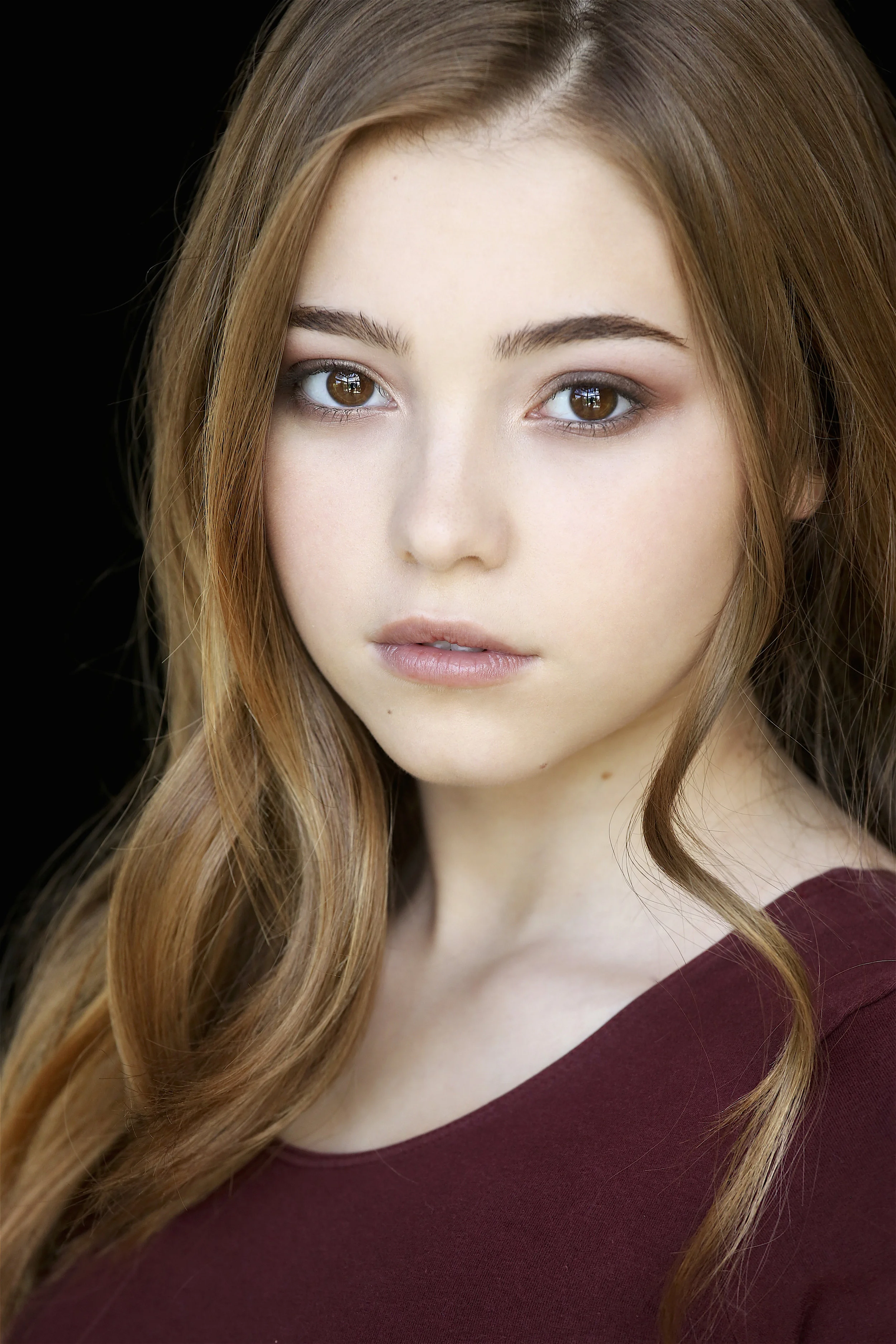 Close-up portrait of a young woman with long, wavy auburn hair, light skin, and brown eyes, wearing a dark red top, against a black background.