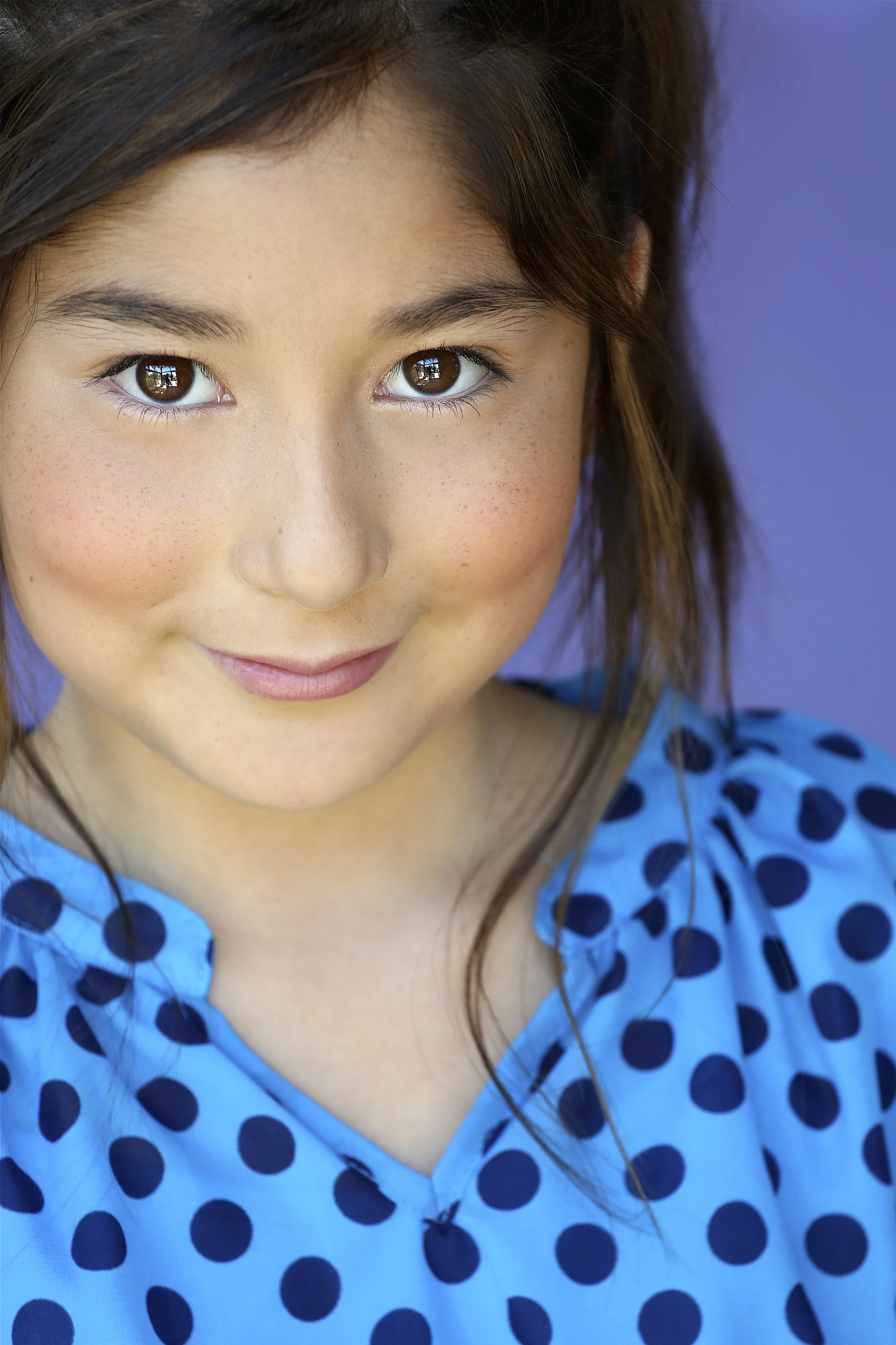 Close-up of a young girl with brown hair and freckles, smiling, wearing a blue dress with dark blue polka dots, against a purple background.