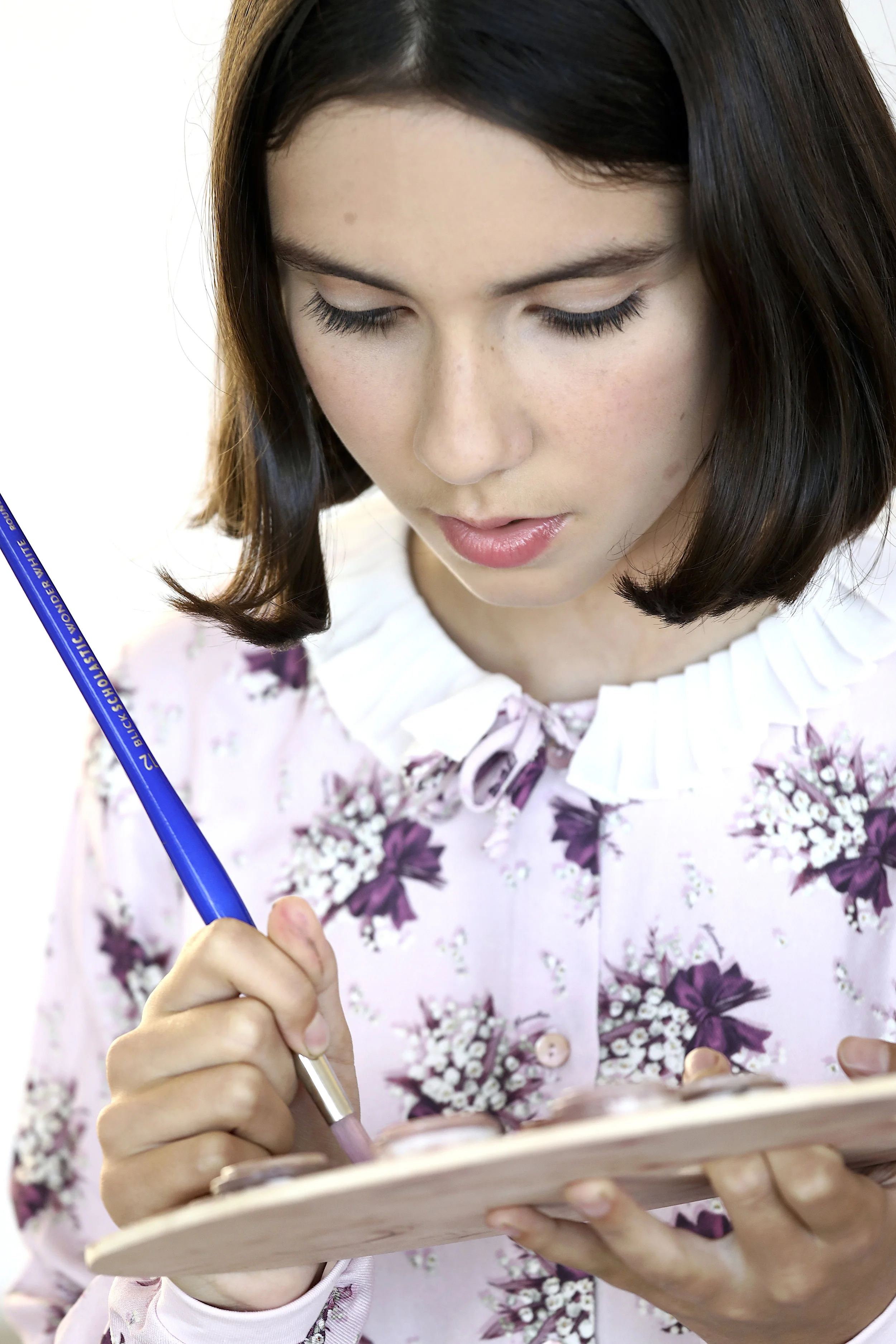 A young woman with dark hair, wearing a pink floral blouse, is holding a small wooden palette and using a brush to paint intricate designs.