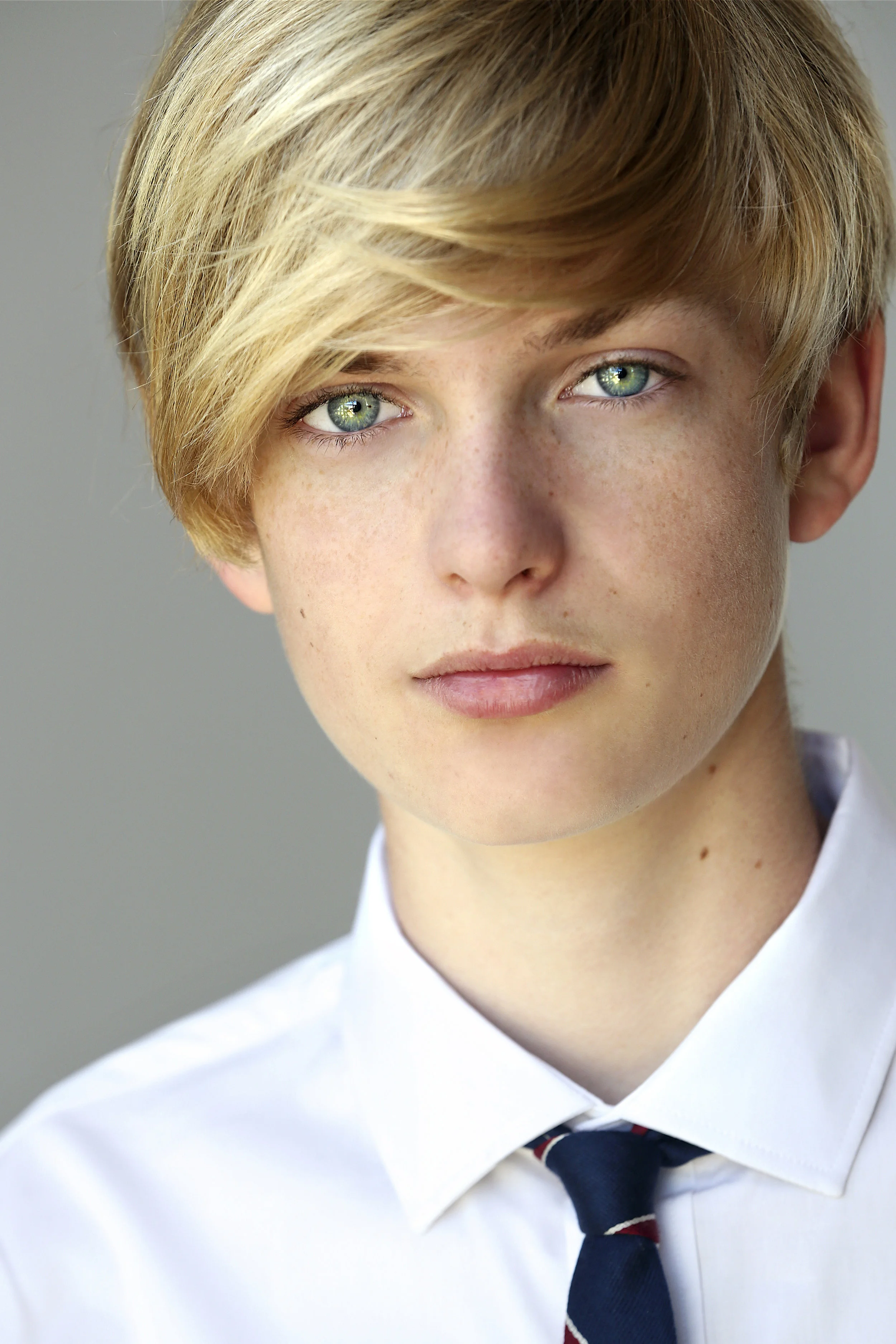 Close-up of a young person with short blonde hair, green eyes, wearing a white shirt and a dark tie.