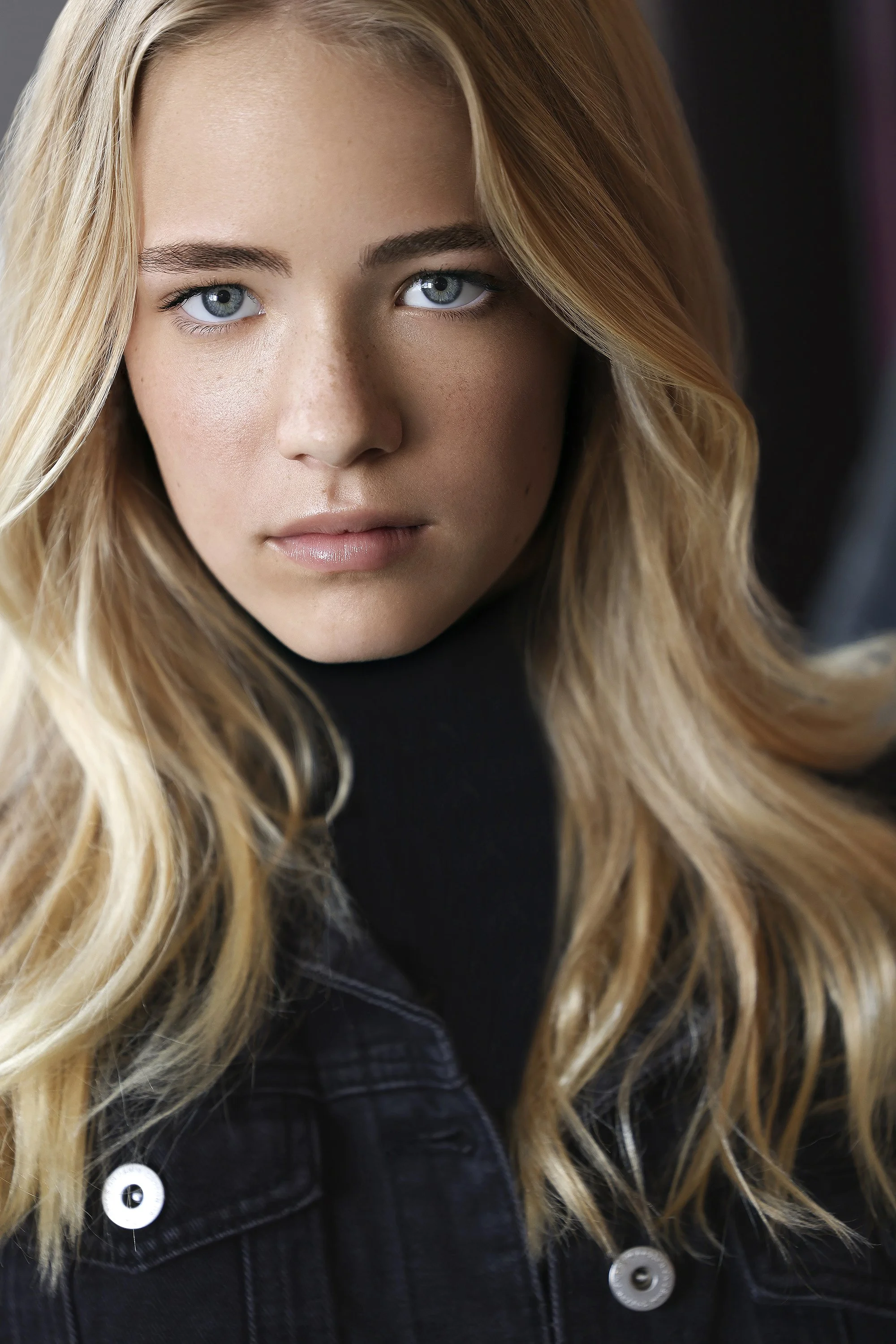 Close-up of a young woman with blonde wavy hair, blue eyes, and light makeup, wearing a black jacket.
