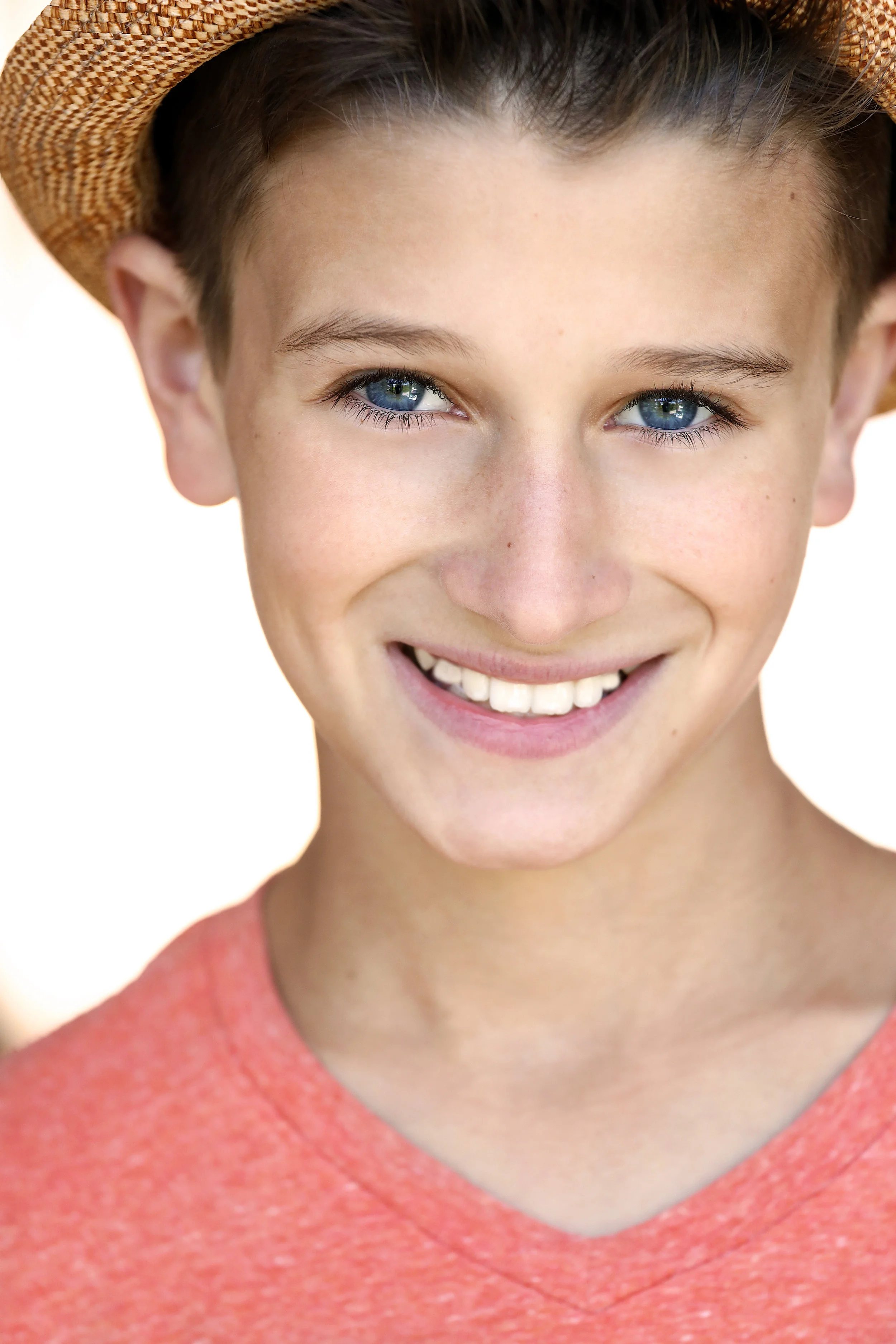 Close-up of a smiling young boy with blue eyes wearing a straw hat and a coral-colored shirt.