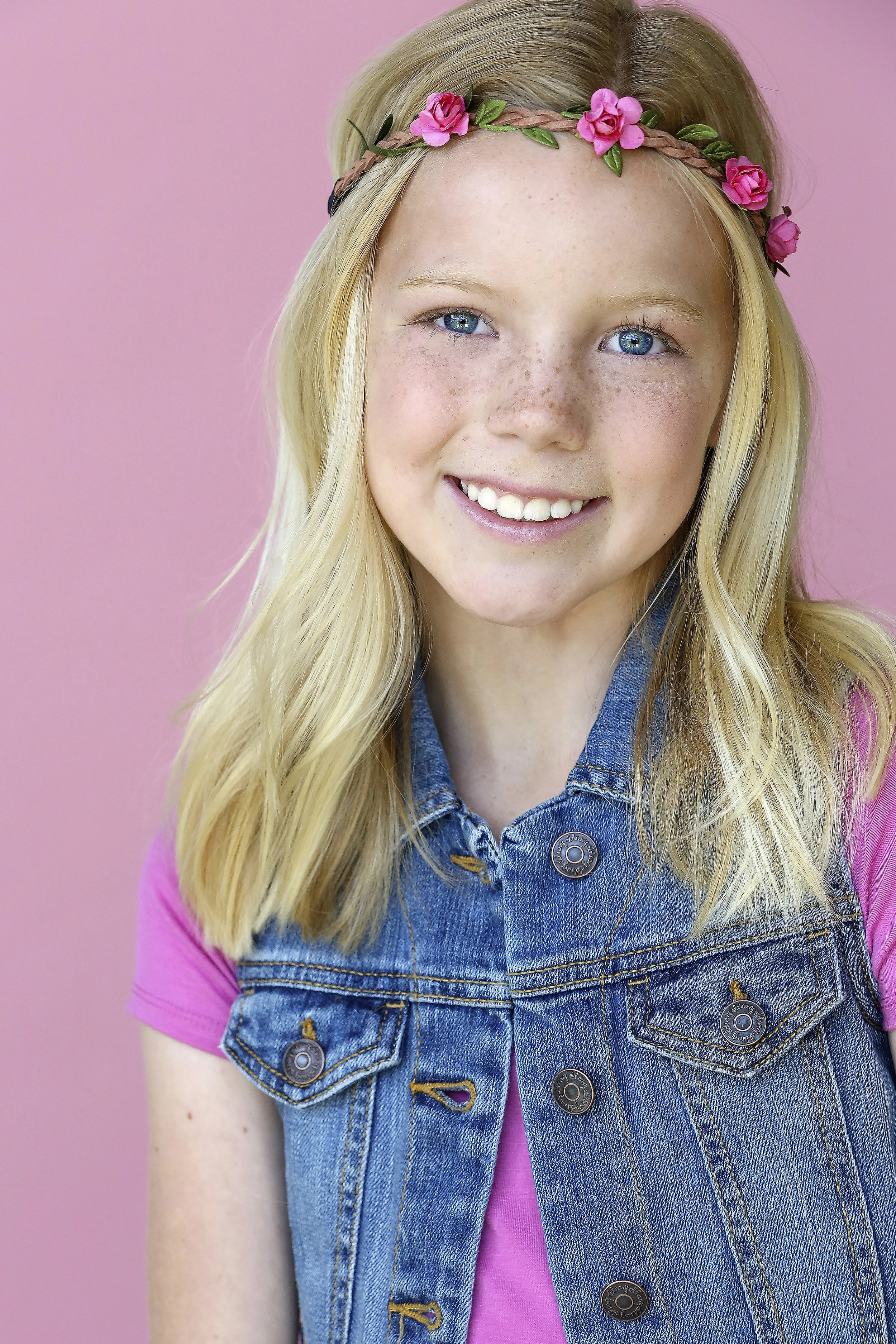 A smiling blonde girl wearing a pink shirt and denim vest, with a flower crown made of pink flowers and green leaves against a pink background.