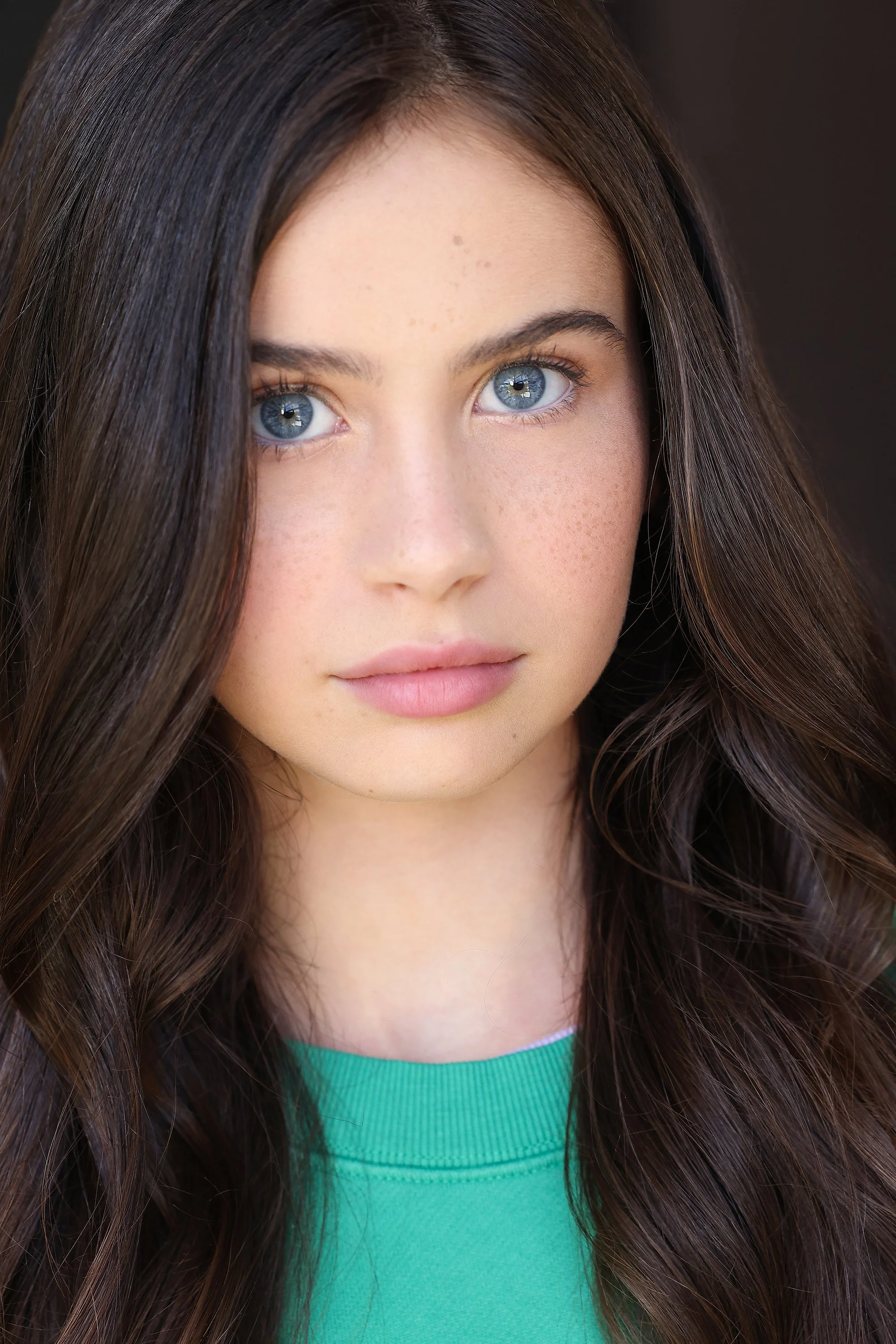 A young woman with long, dark brown hair and blue eyes, wearing a green top, looking directly at the camera.