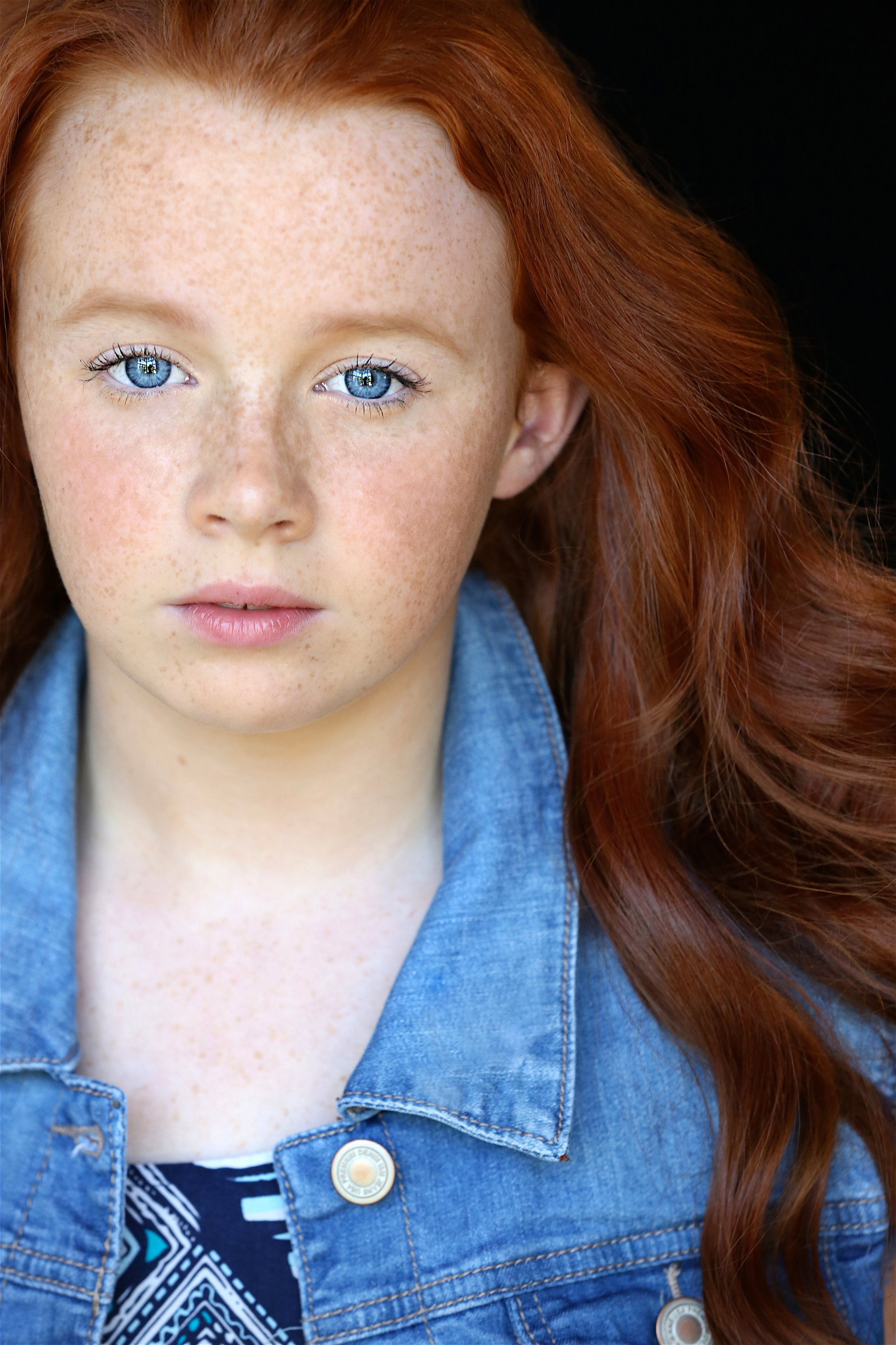 A young girl with bright blue eyes, freckles, and long red hair wearing a denim jacket and a patterned top.