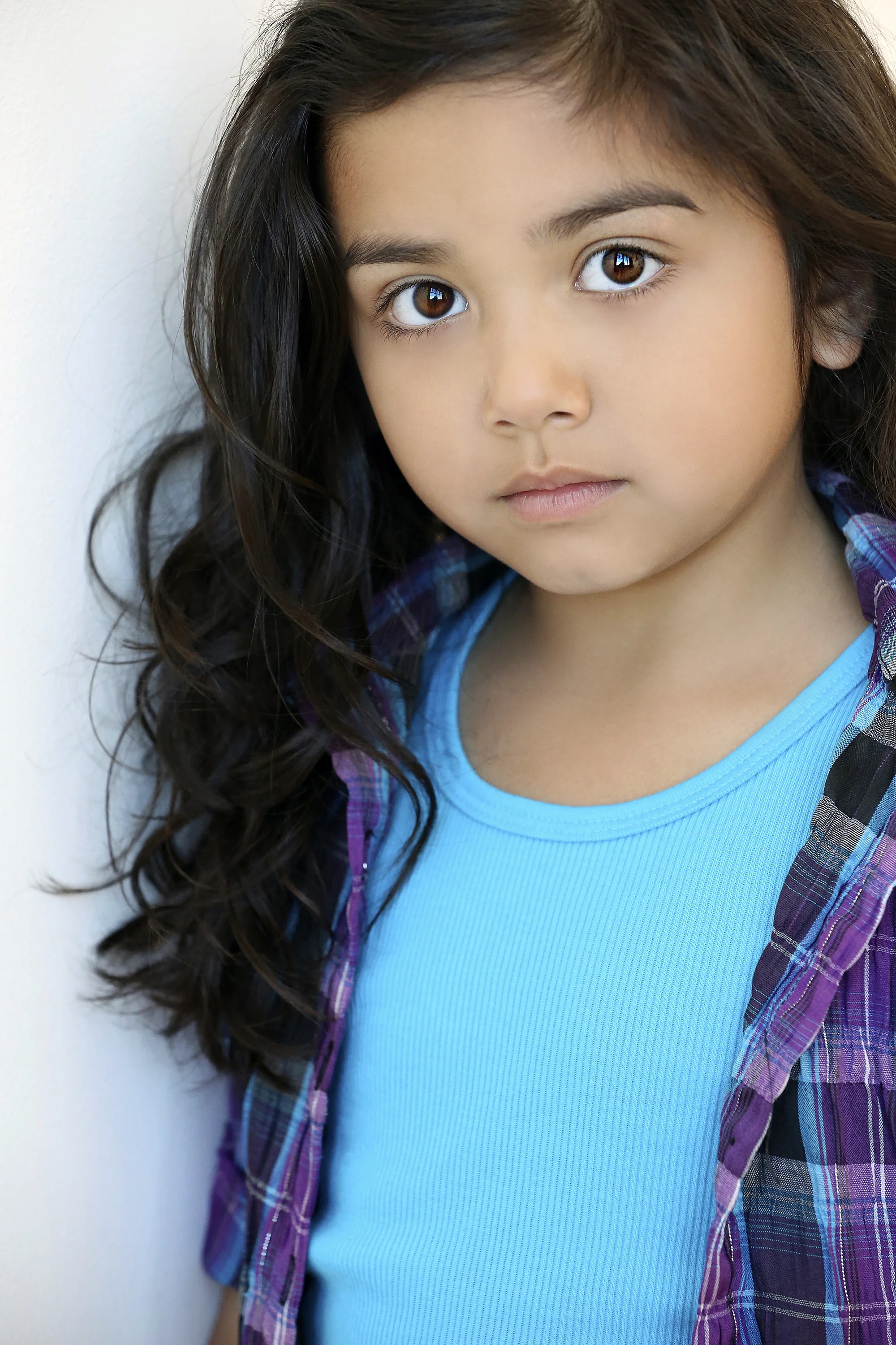 A young girl with long dark hair, wearing a bright blue shirt and a purple plaid shirt, standing against a white wall, looking directly at the camera with a neutral expression.
