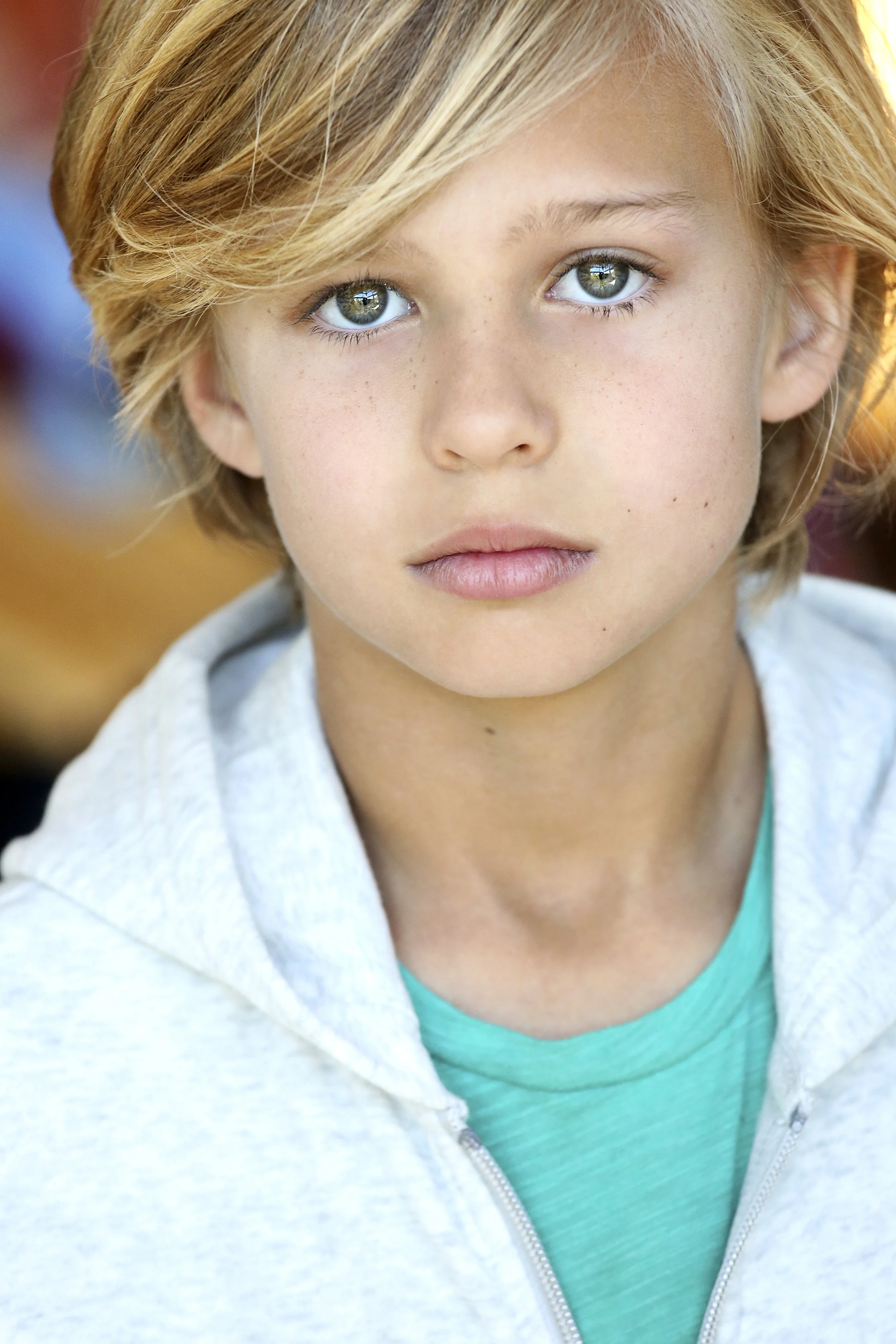 A young boy with blonde hair and blue eyes looking directly at the camera, wearing a light gray hoodie and a teal shirt underneath.