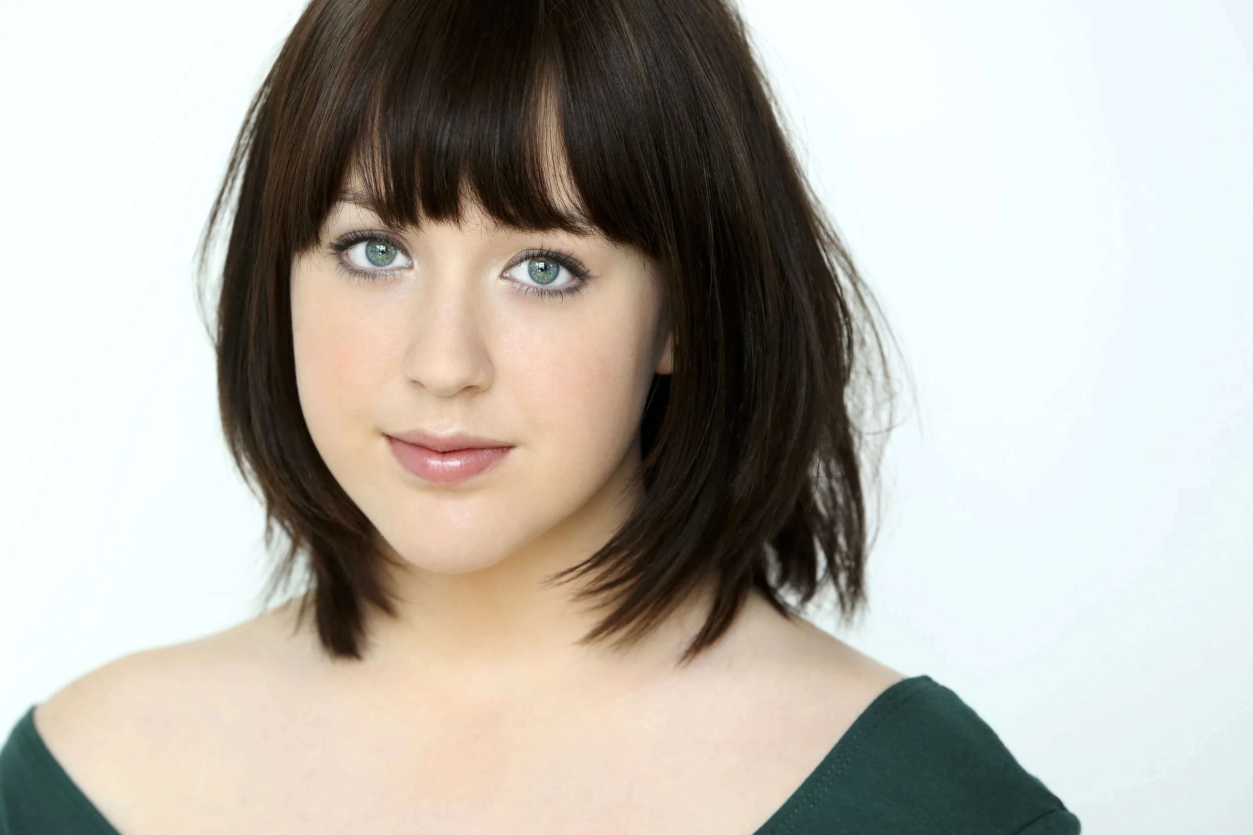 Close-up portrait of a young woman with shoulder-length brown hair, green eyes, and fair skin, wearing a dark green top.