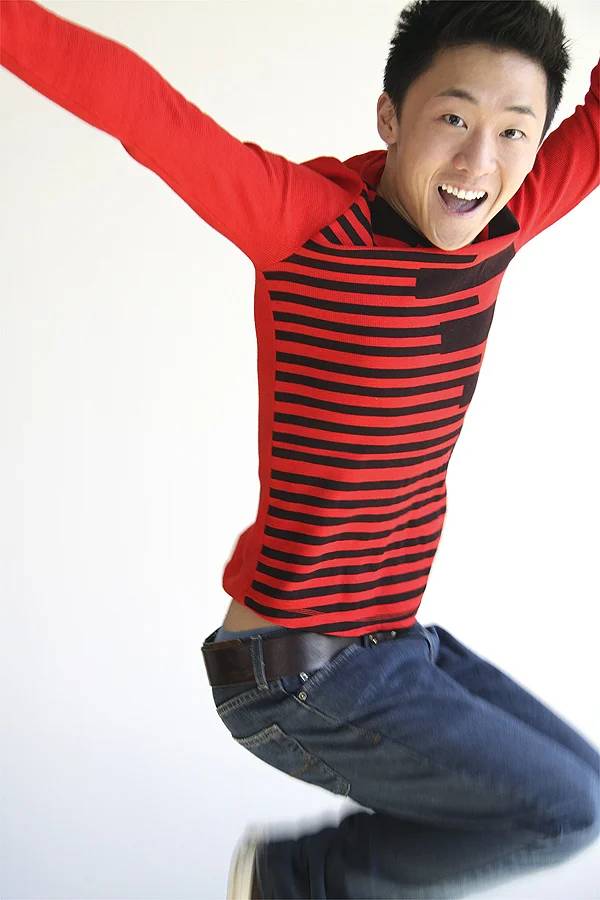 A young man jumping in the air with arms raised, smiling, wearing a red and black striped long-sleeve shirt and blue jeans.