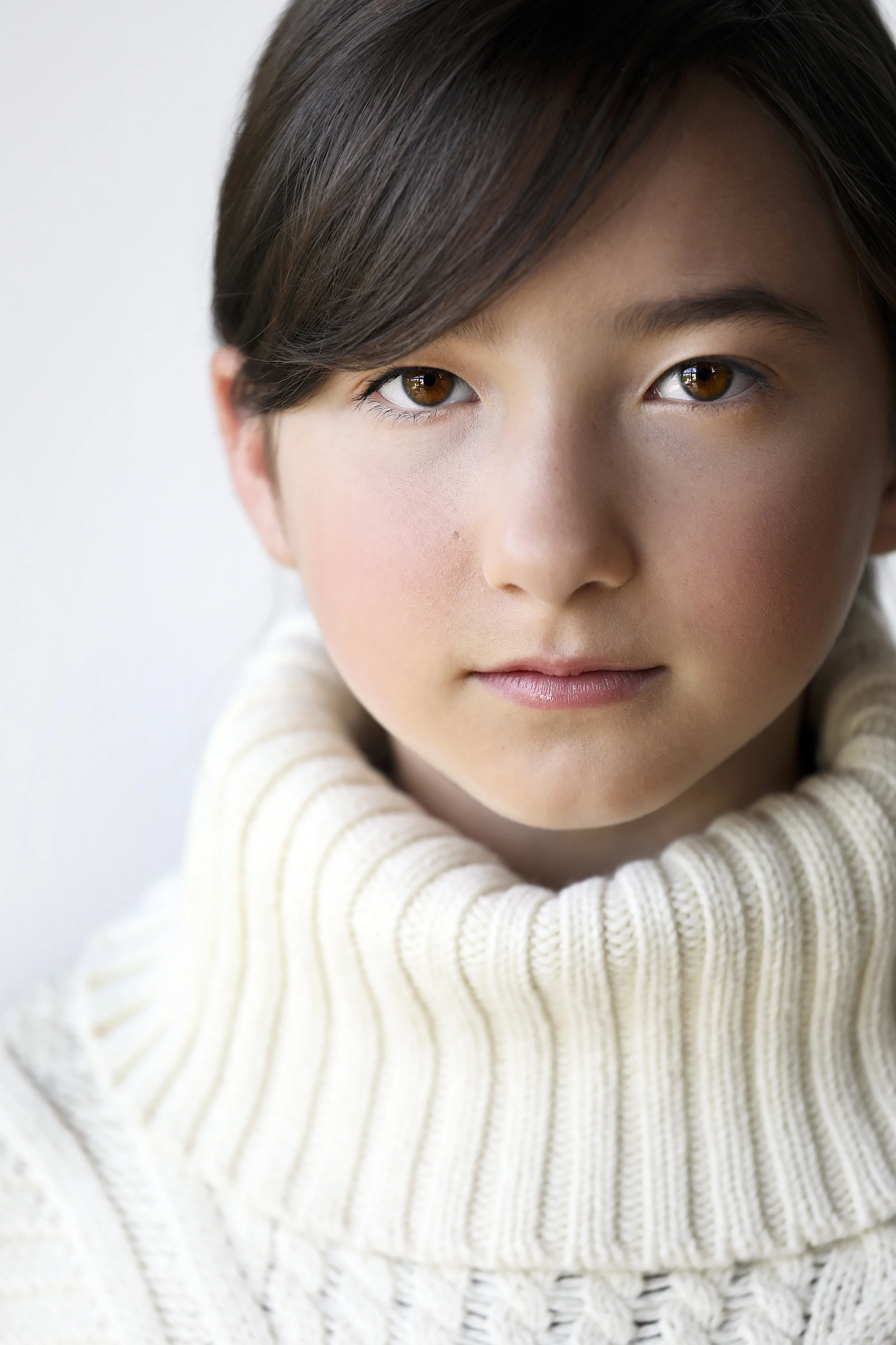 Close-up portrait of a young woman with brown hair, brown eyes, and a neutral expression, wearing a white turtleneck sweater.