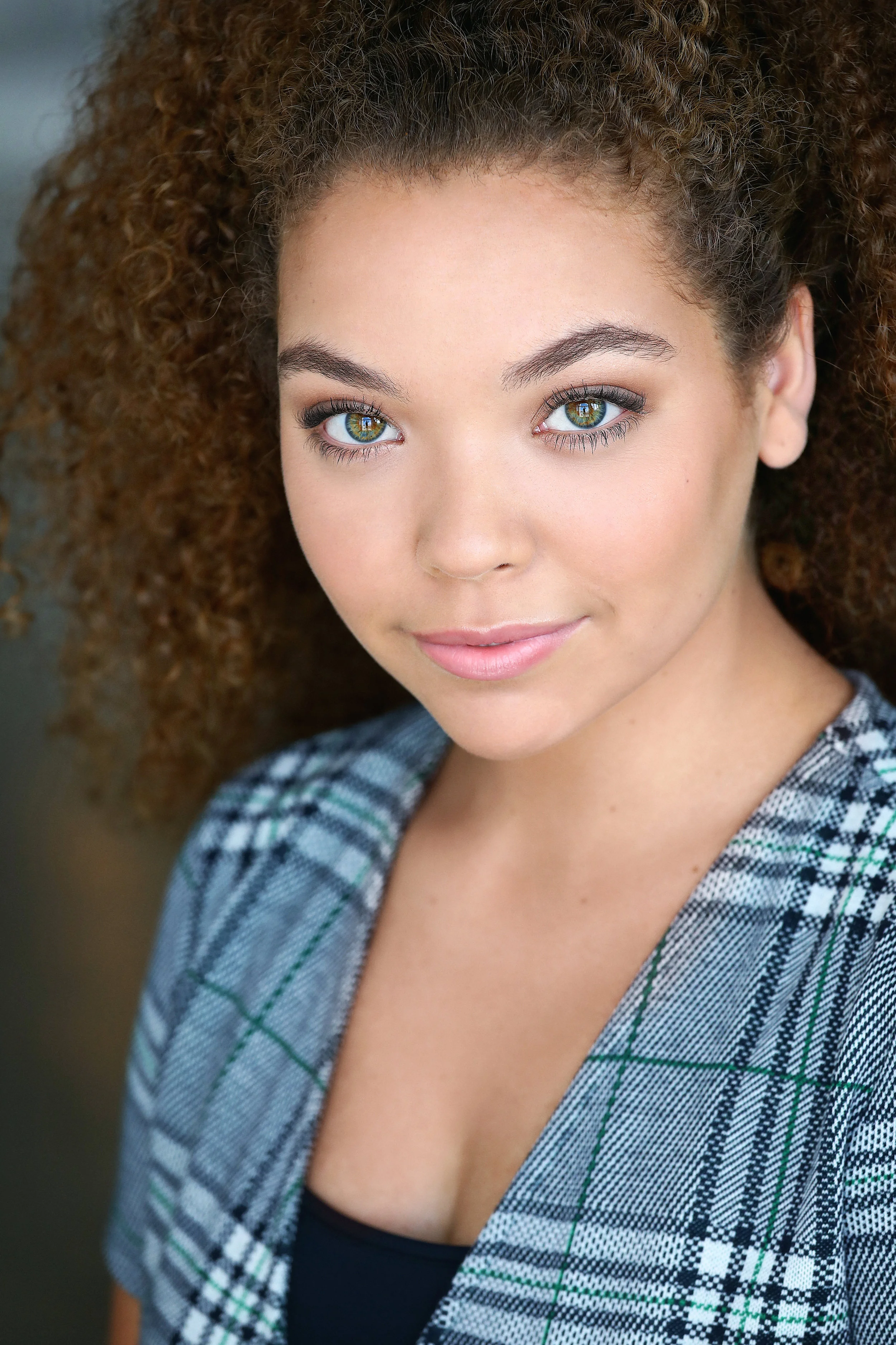 Close-up of a young woman with curly brown hair, blue eyes, and a light makeup look, wearing a plaid blazer over a black top.
