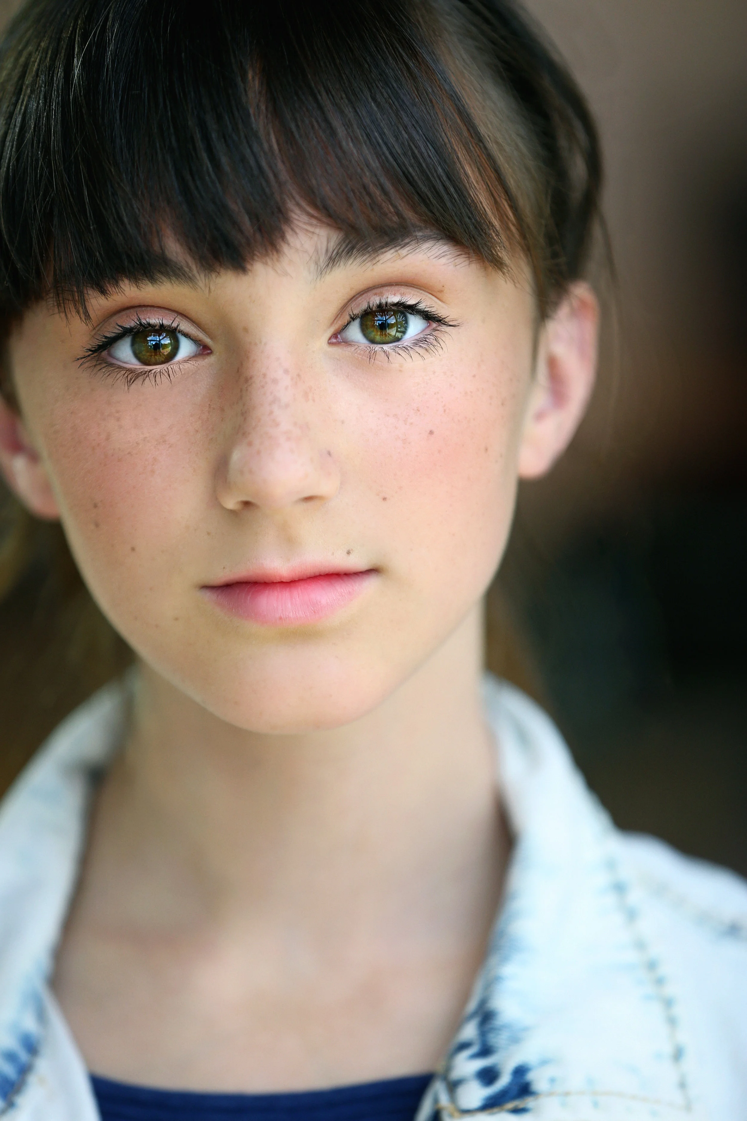 Close-up of a young girl with brown hair, green eyes, and freckled face, looking directly at the camera.