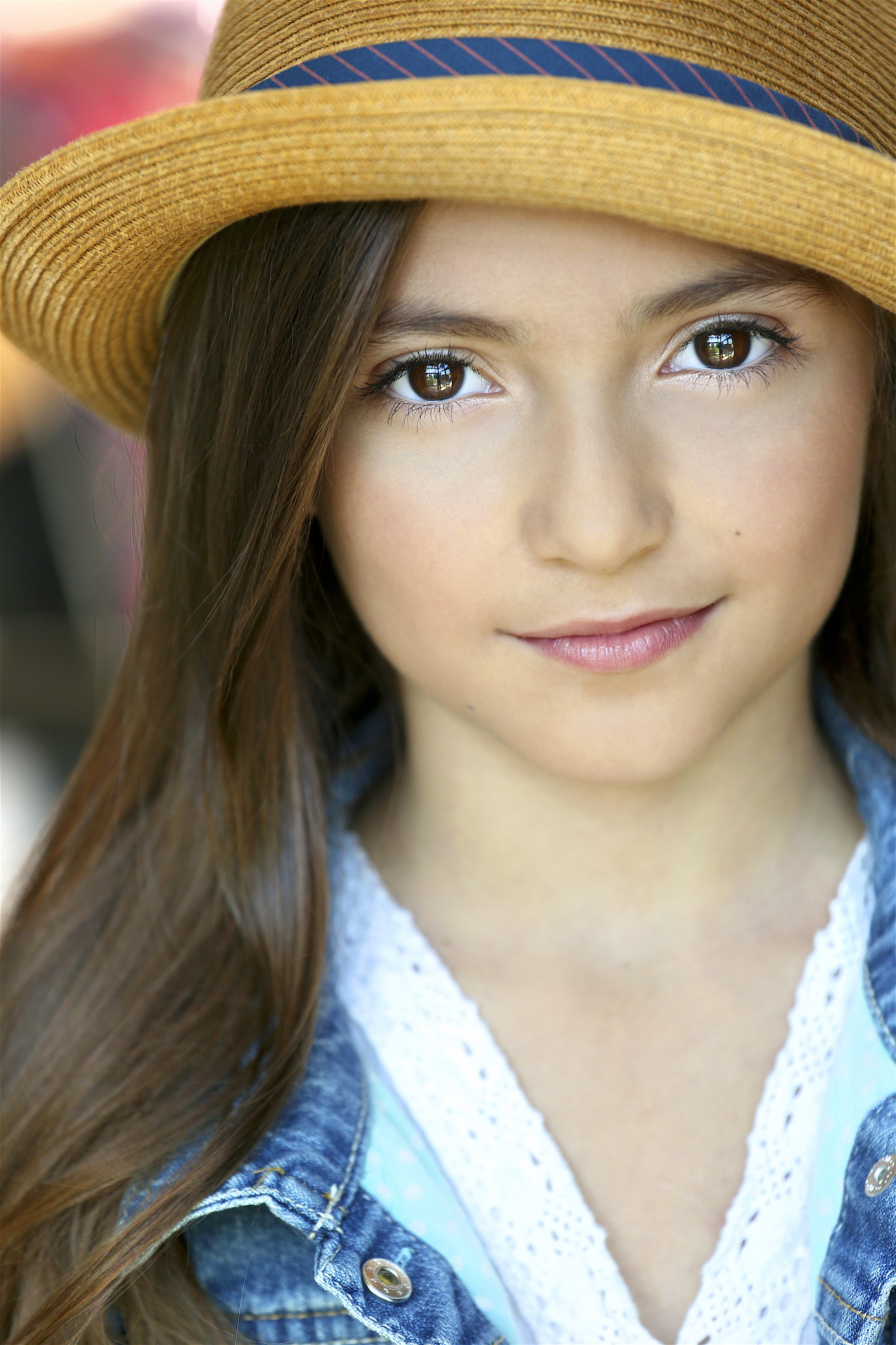 Close-up of a young woman with long brown hair wearing a straw hat, denim jacket, and a white blouse, smiling softly.
