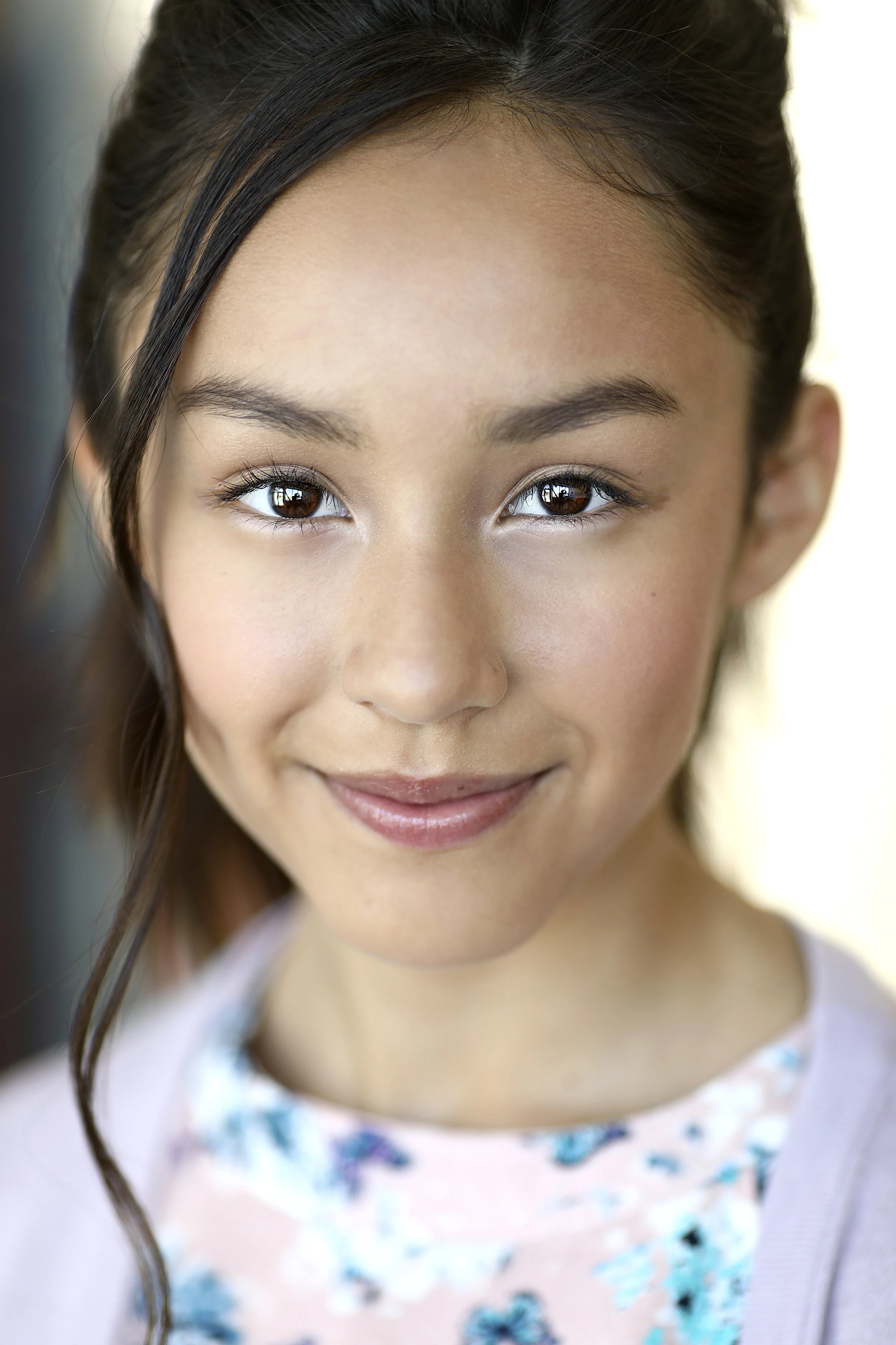 Close-up of a young girl with brown eyes, smiling, with brunette hair styled with loose waves.