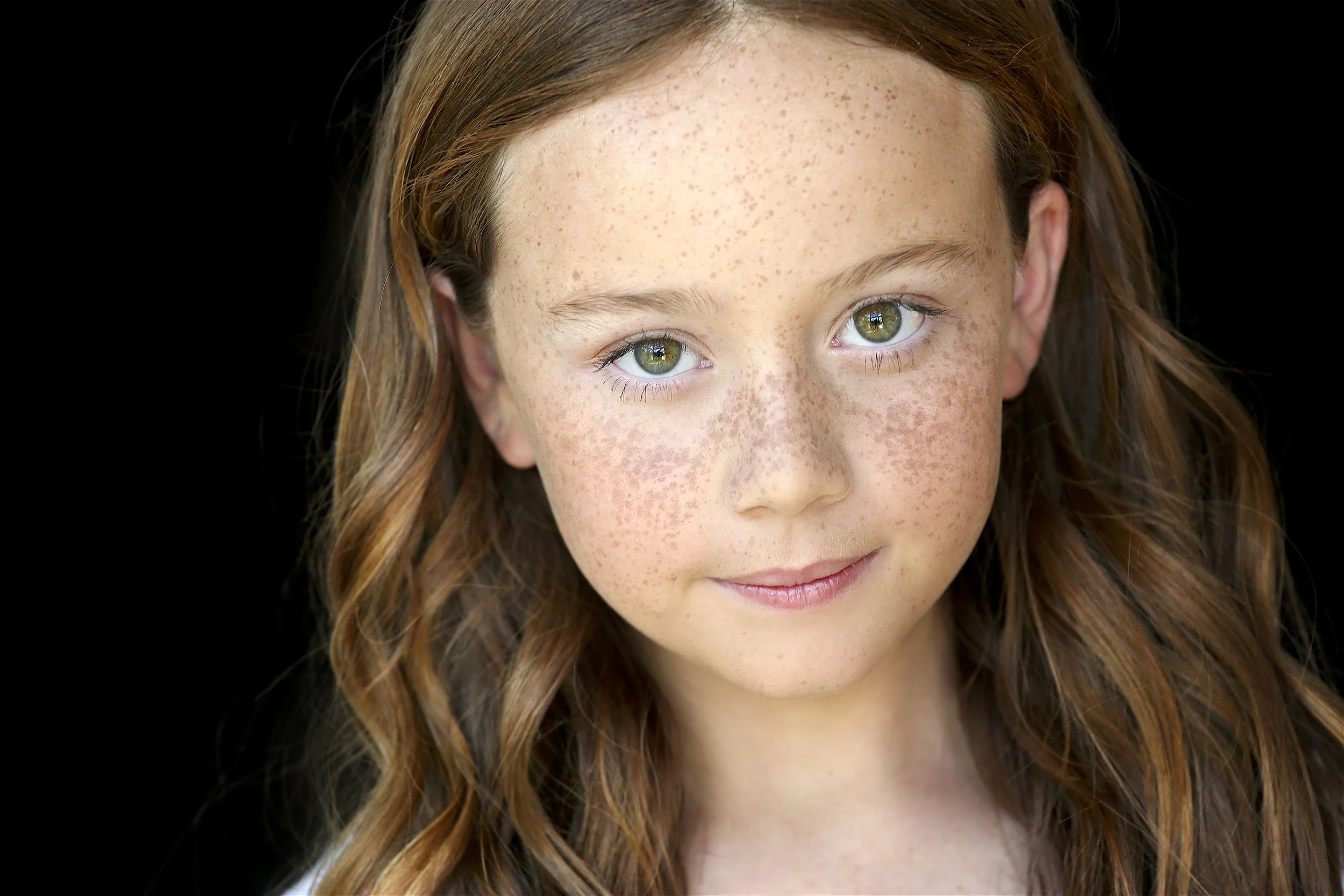 Close-up of a young girl with red hair and freckles, smiling softly, against a black background.