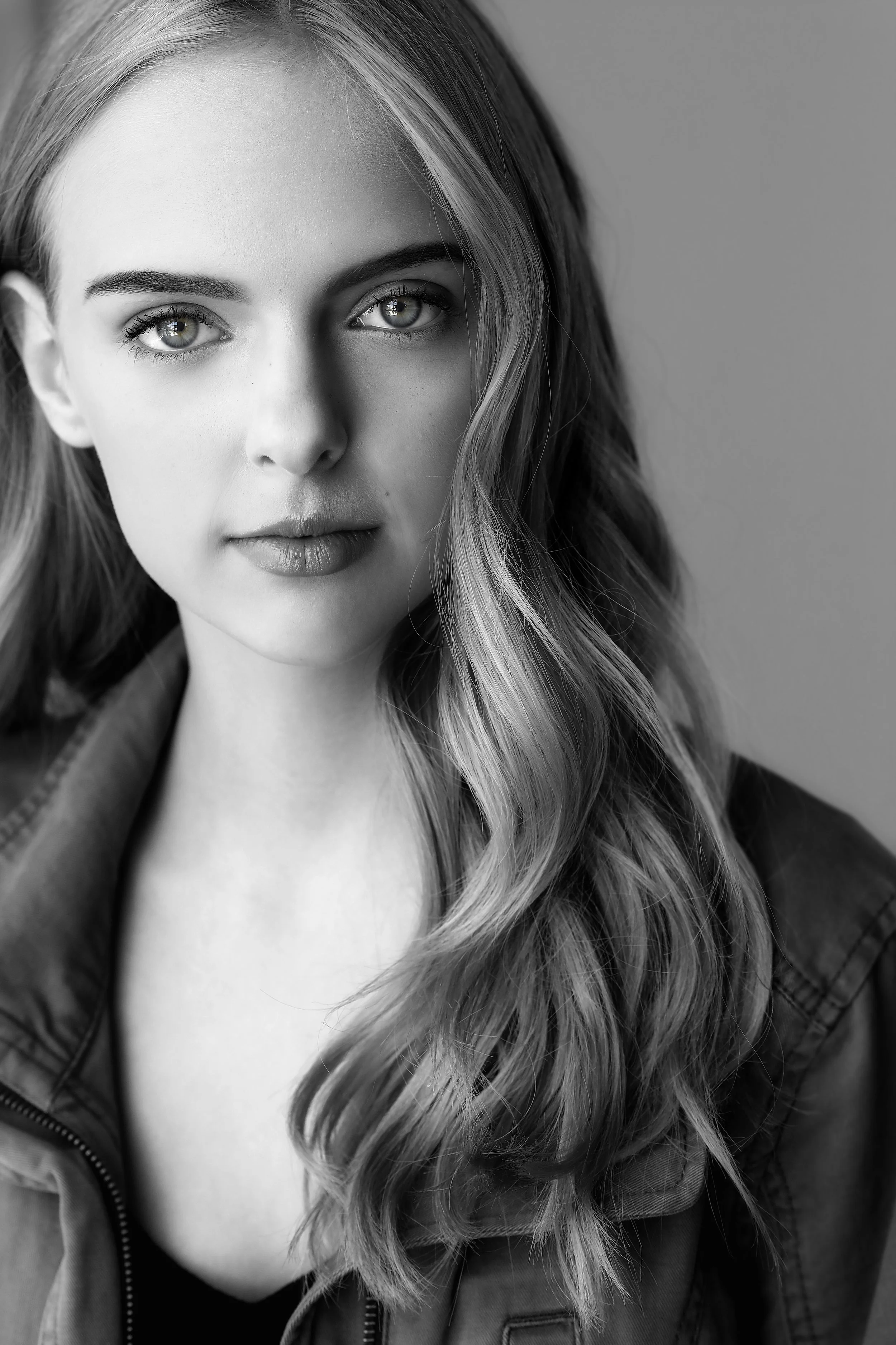 Black and white portrait of a young woman with wavy hair, gazing at the camera with a serious expression, wearing a jacket.