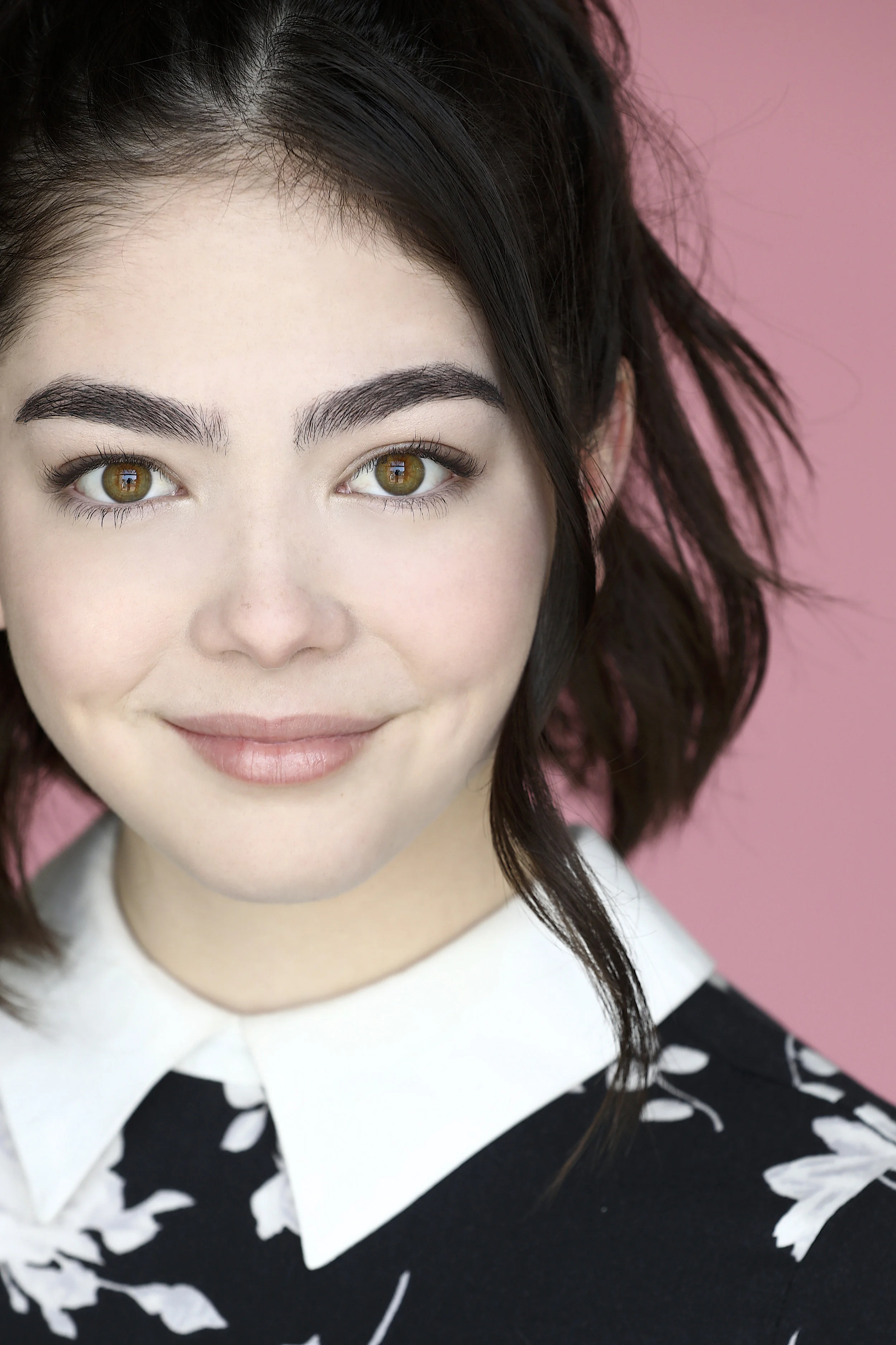 Close-up portrait of a smiling young woman with dark hair, hazel eyes, and fair skin, wearing a black and white floral top with a white collar, against a pink background.