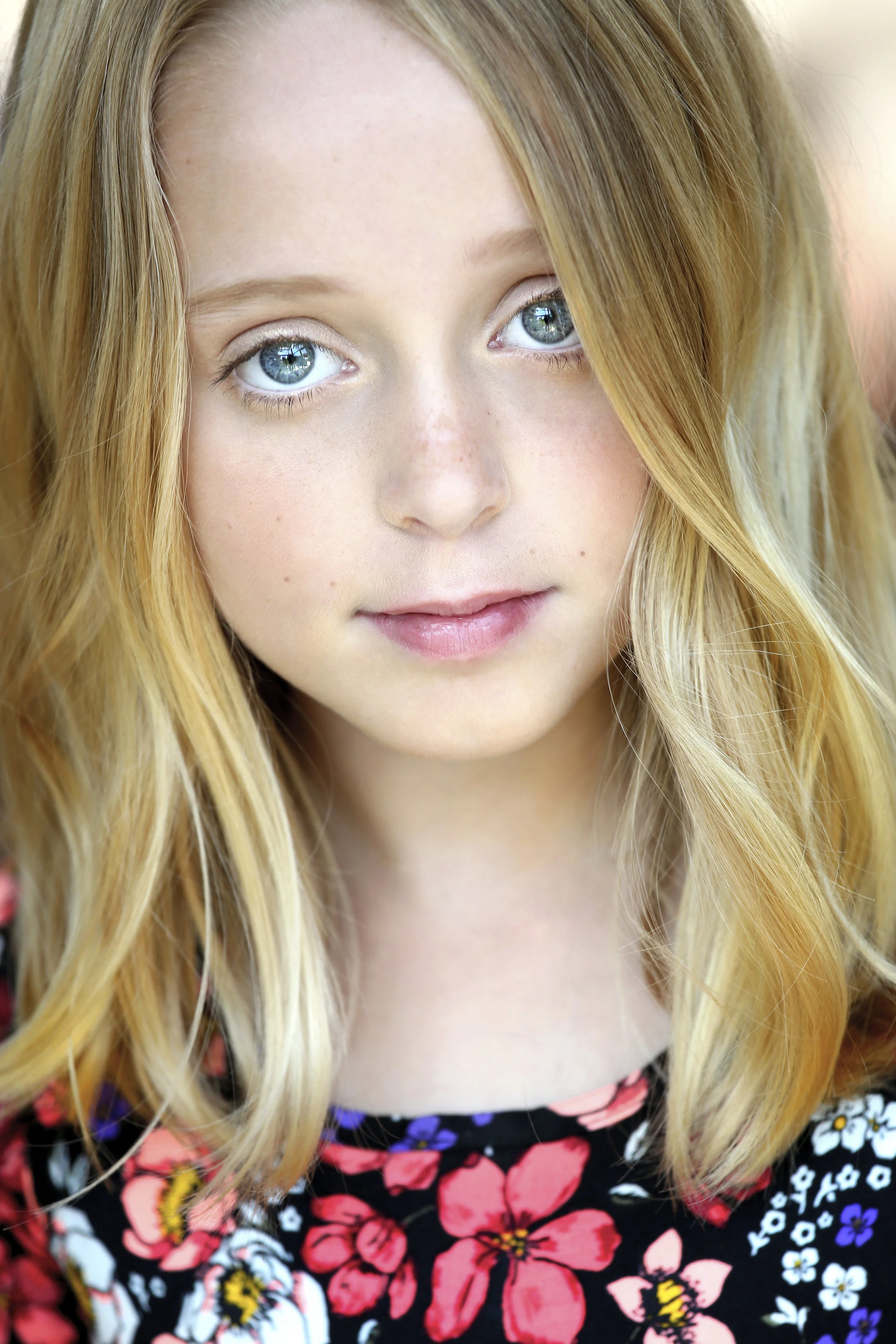 Close-up of a young girl with blonde hair, blue eyes, and light skin, wearing a black top with colorful floral print.