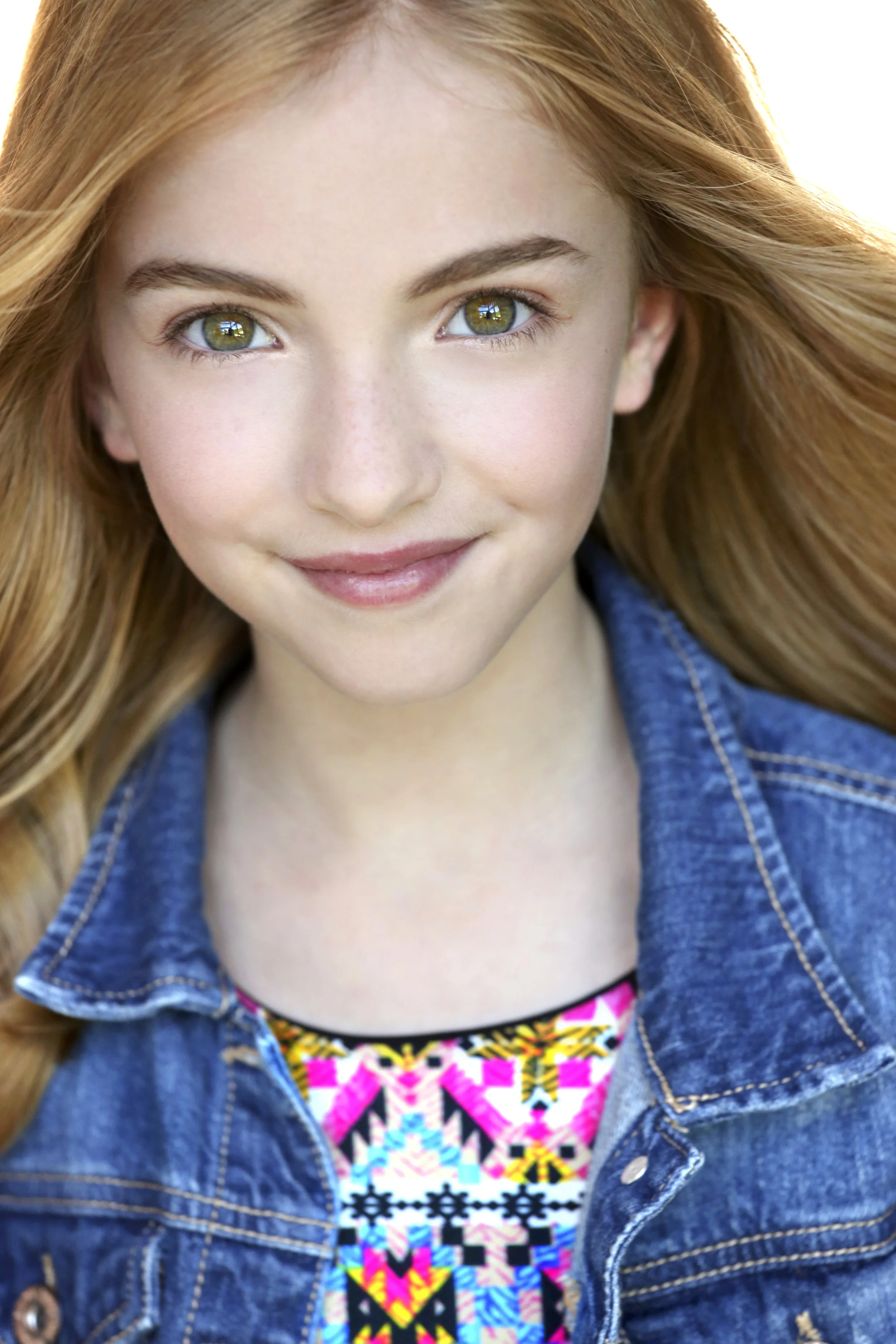 Close-up portrait of a young girl with long red hair, green eyes, freckles, wearing a denim jacket and a colorful patterned top, smiling gently.