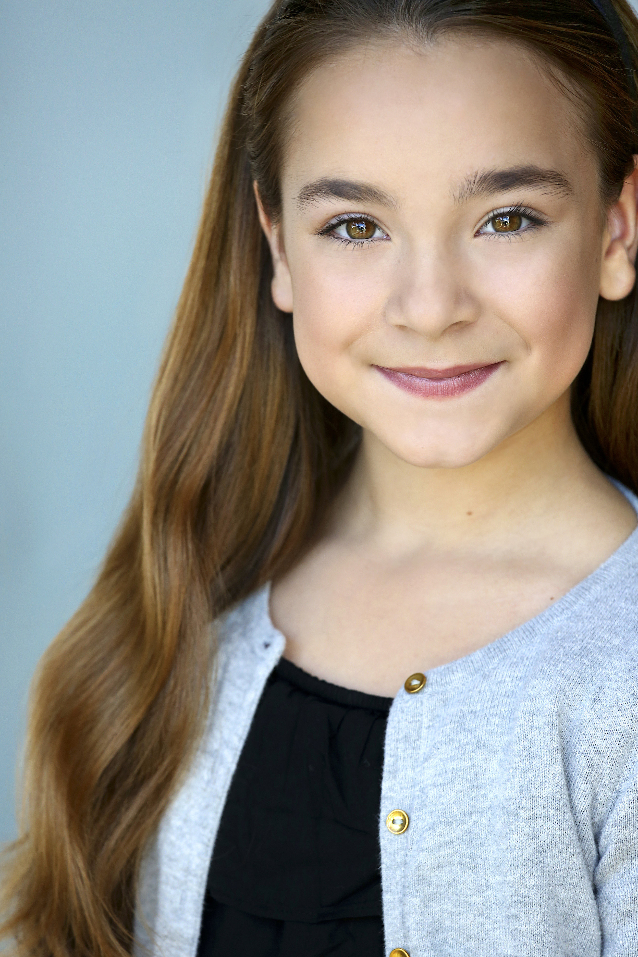 Close-up portrait of a young girl with long, wavy red hair, brown eyes, and a warm smile, wearing a gray cardigan over a black top, standing against a light blue background.
