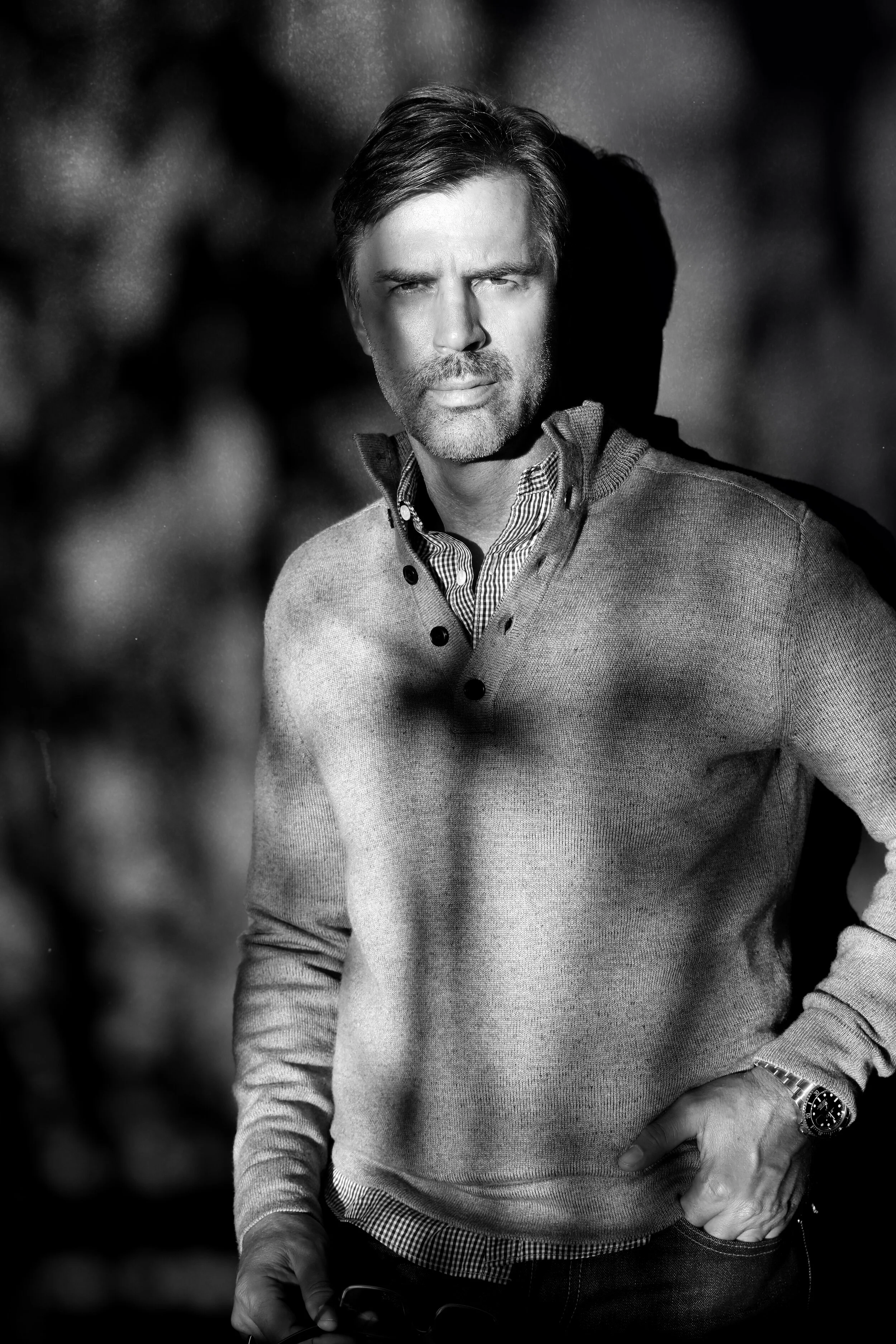 A black and white photo of a middle-aged man with a beard, dressed in a light sweater over a checkered shirt, standing outdoors with a blurred background.