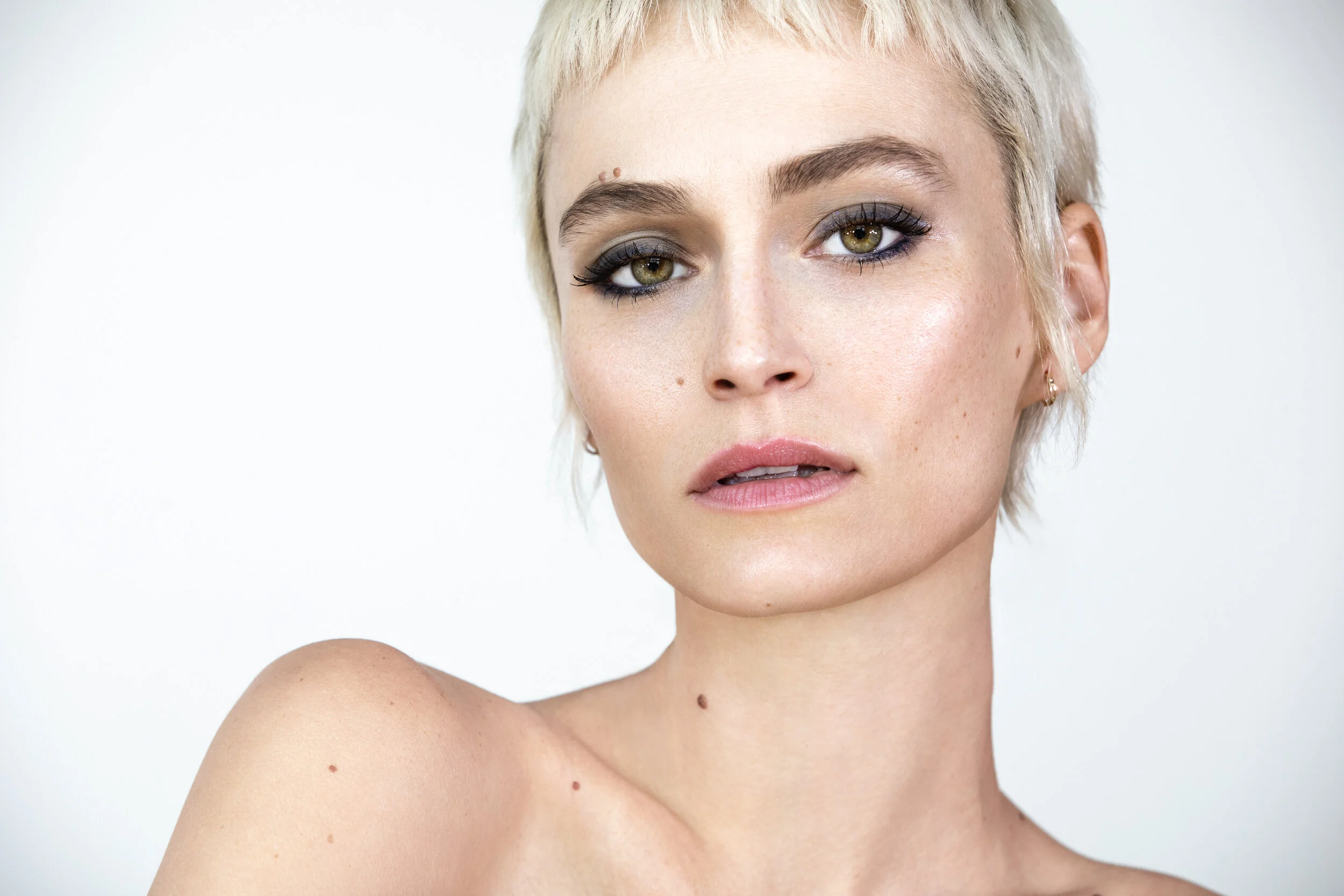 Close-up portrait of a woman with short blonde hair, wearing makeup with bold eyeliner and mascara, and a natural lip color, with fair skin and a few moles visible, against a plain white background. Model photographed by Los Angeles photographer Kenn