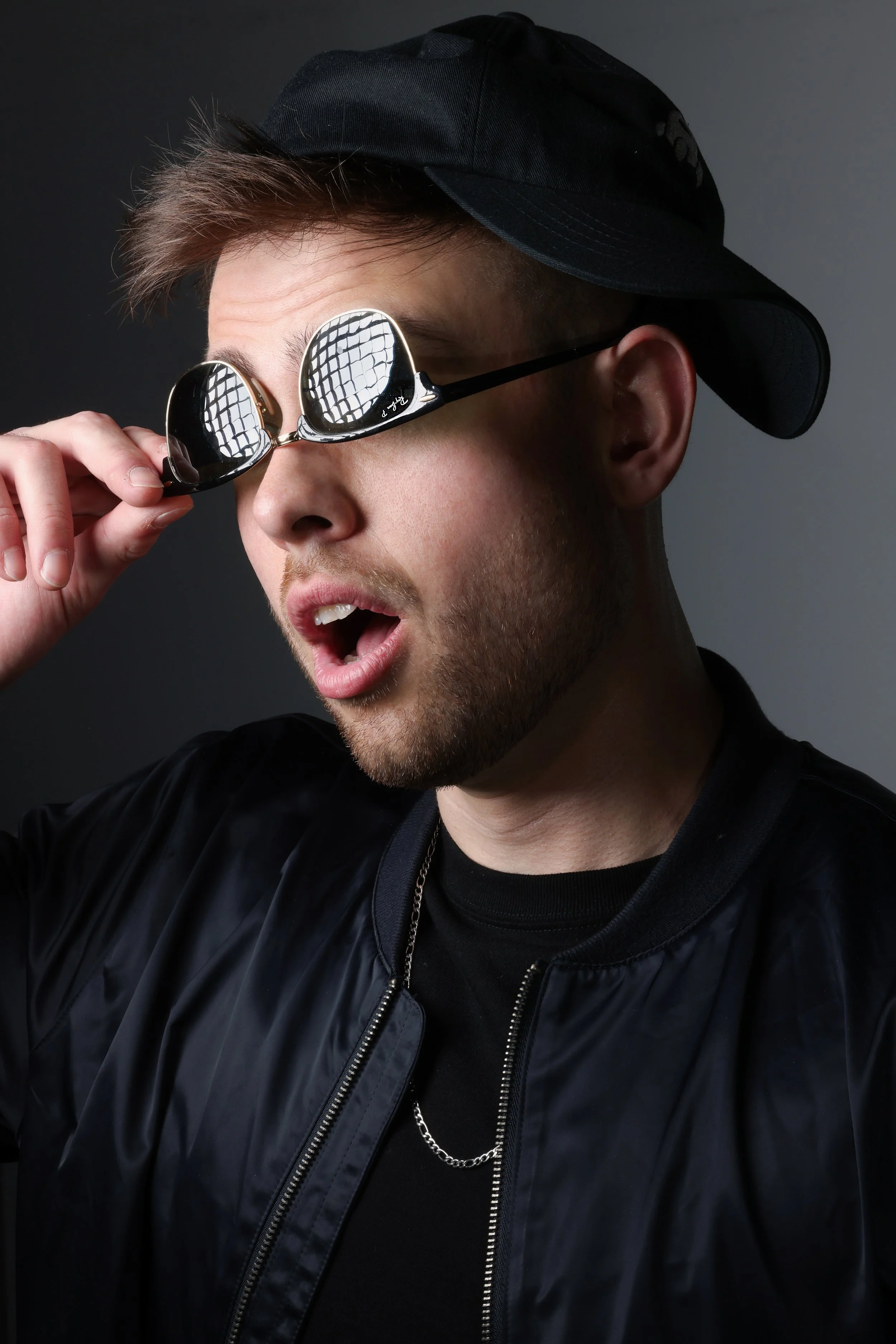 A young man with short hair and a beard wearing sunglasses, a black cap backwards, a black jacket, and a chain necklace, making an expressive surprised or amazed face.