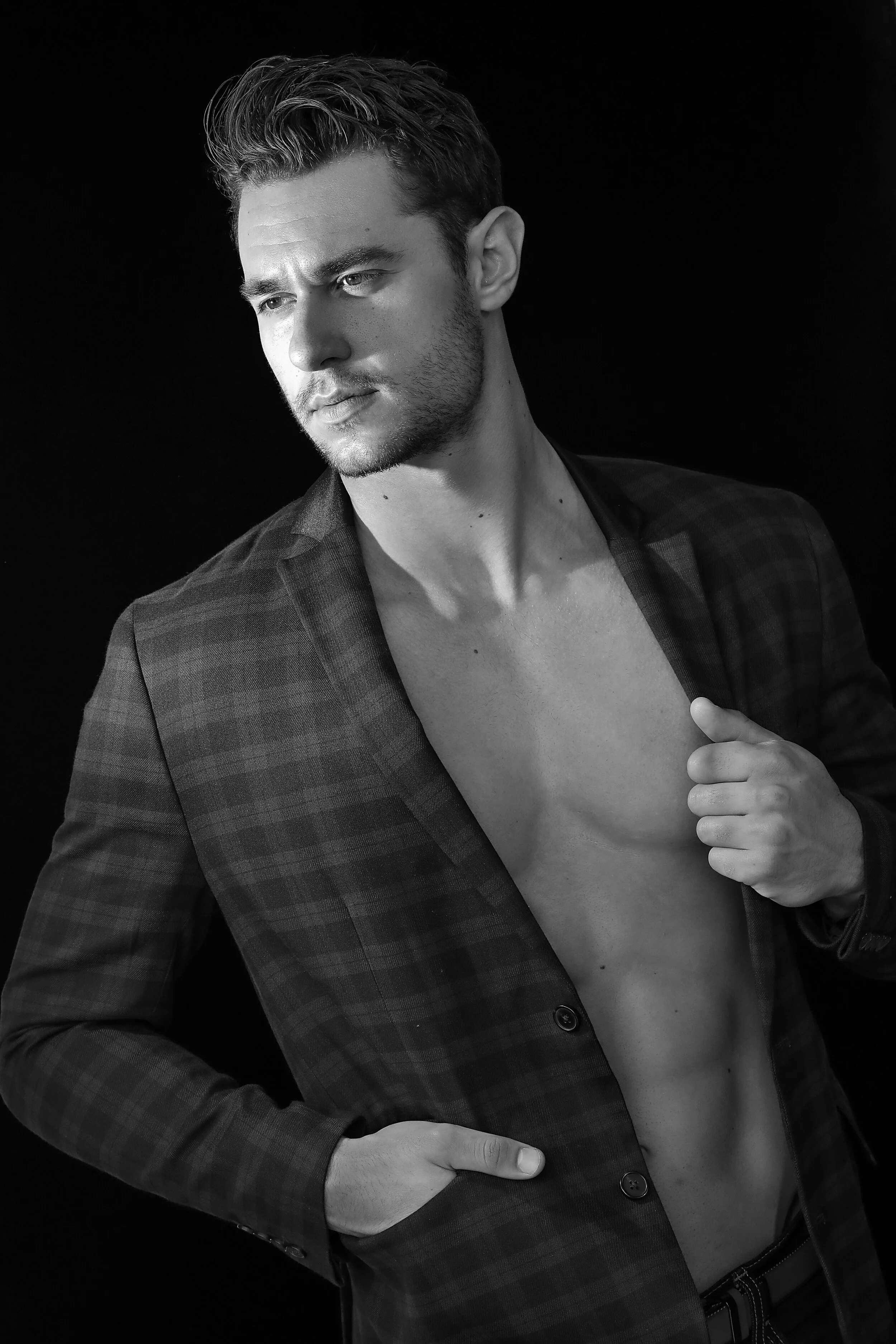 Black and white portrait of a young man with styled hair, wearing a checkered blazer, partially open to show his chest, against a dark background.