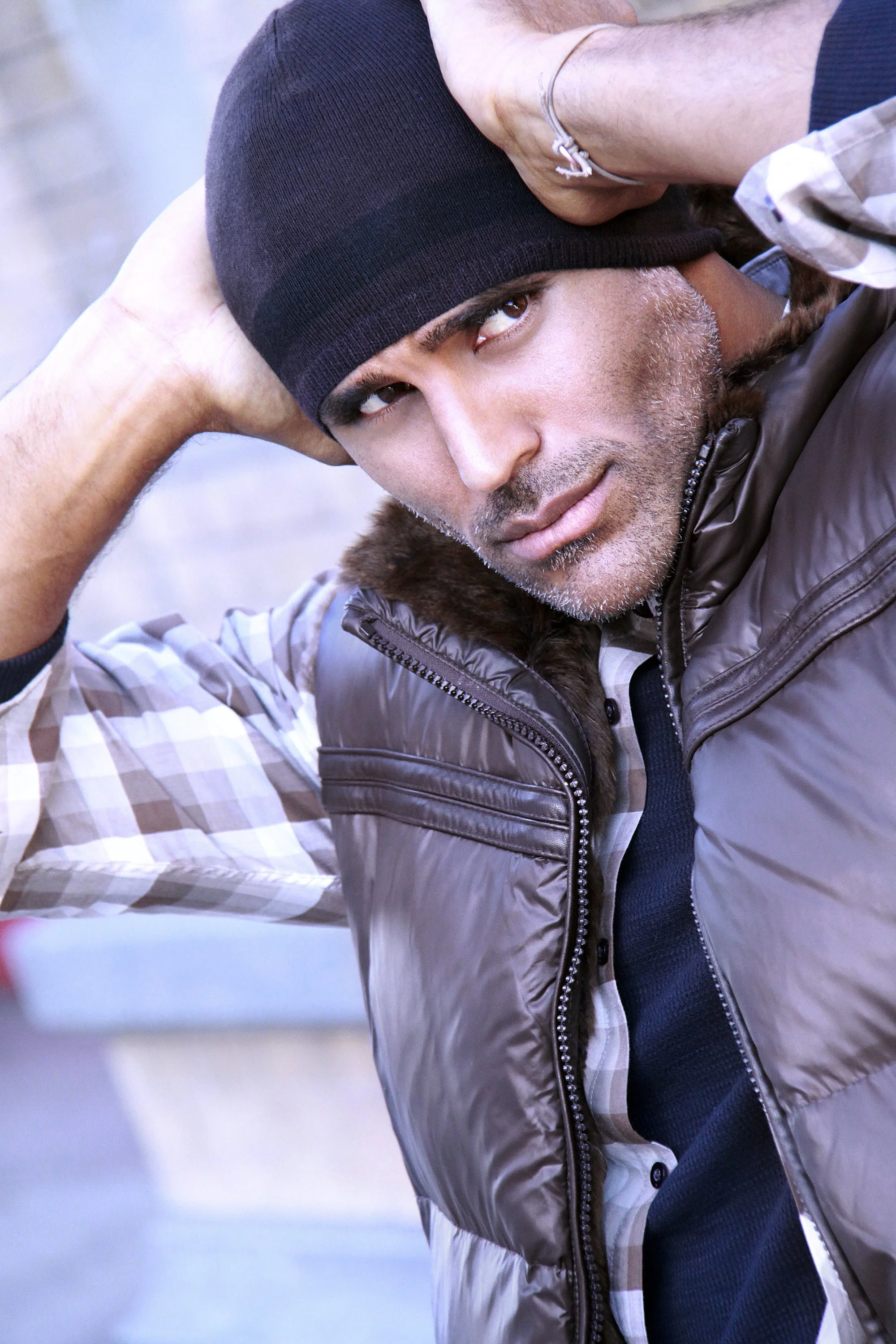 A man wearing a black beanie, brown puffer vest, and plaid shirt, posing outdoors with one hand behind his head and looking at the camera.