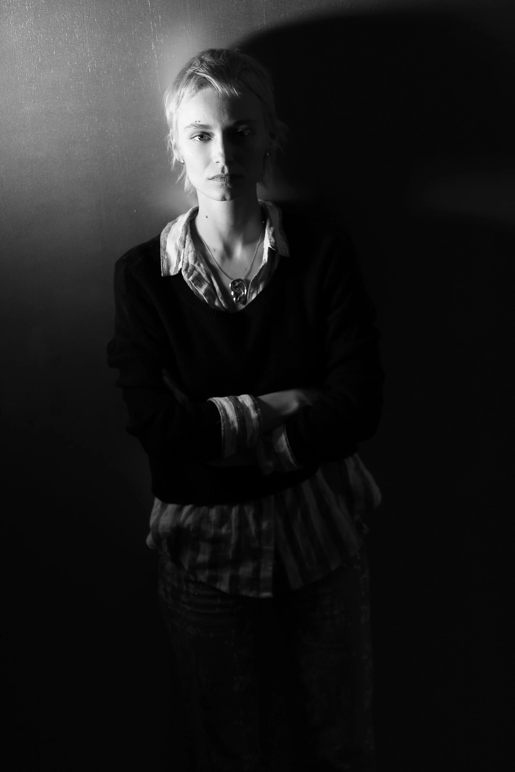 A black and white photo of a young woman with short hair standing against a wall with shadow on the right side, wearing a layered shirt and sweater, with arms crossed.