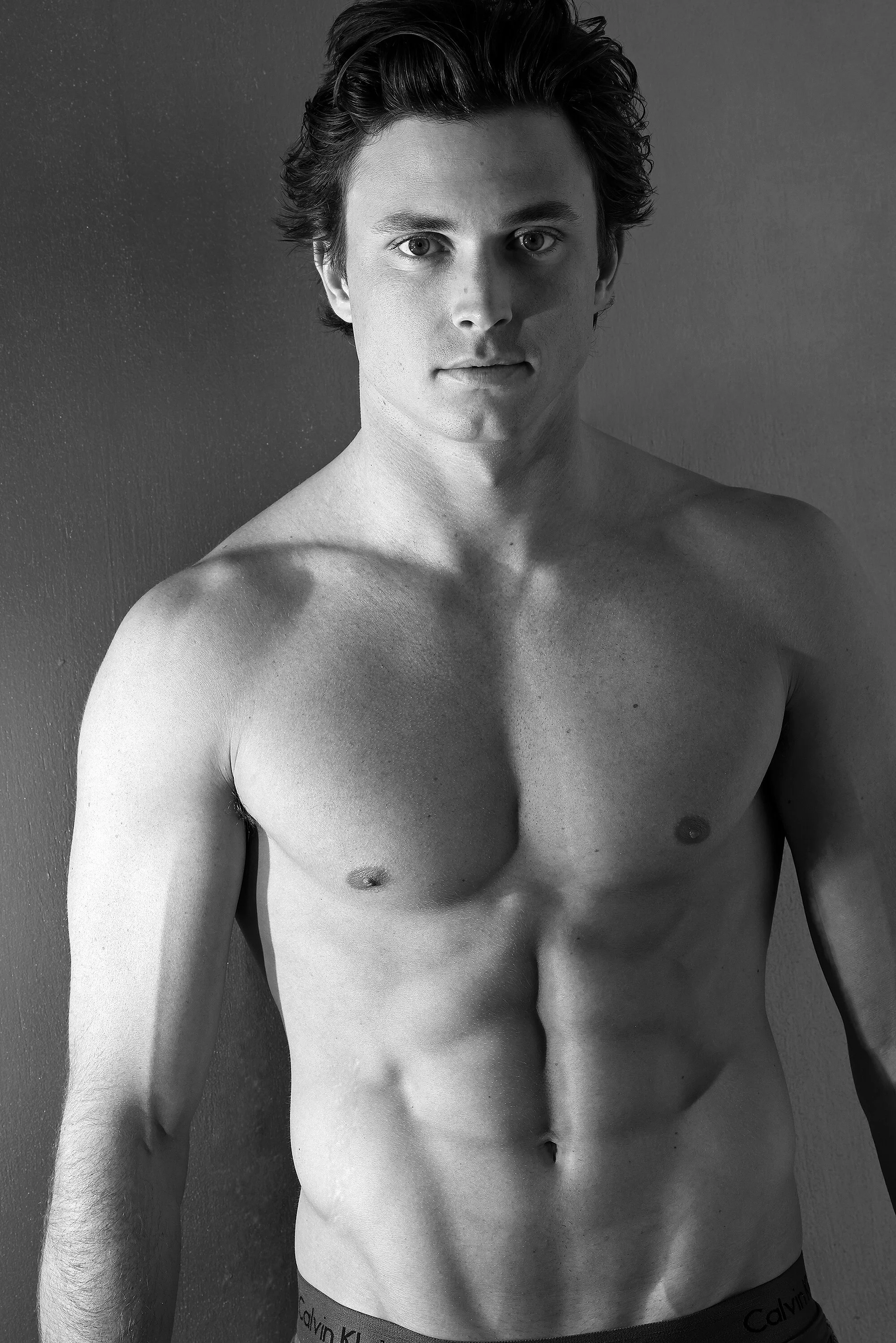 Black and white photo of a shirtless young man with dark, wavy hair looking directly at the camera, standing against a plain wall.