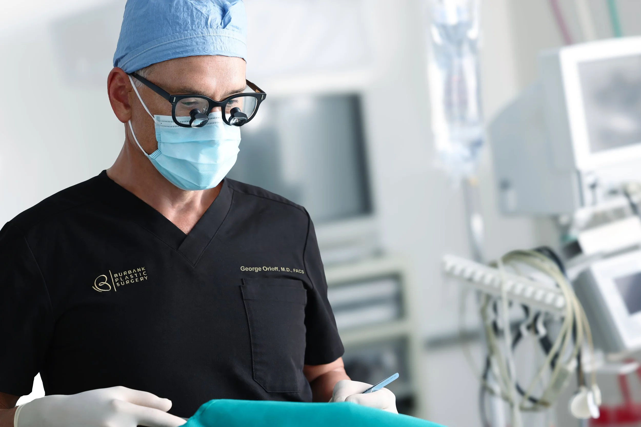 A surgeon wearing magnifying glasses, scrubs, a mask, and gloves, working in an operating room.