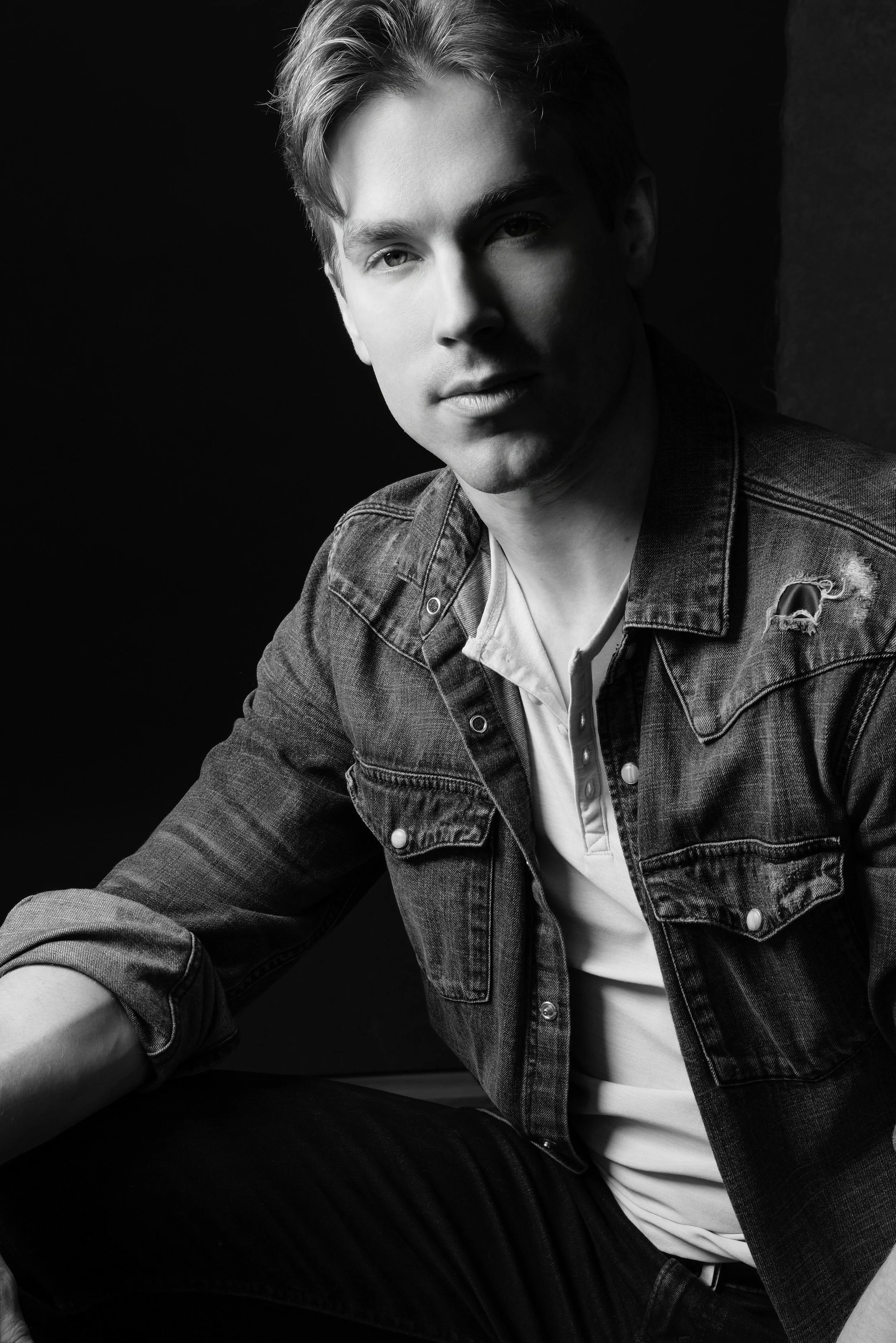 Black and white portrait of a young man with light hair, wearing a denim jacket over a white shirt, sitting against a dark background.