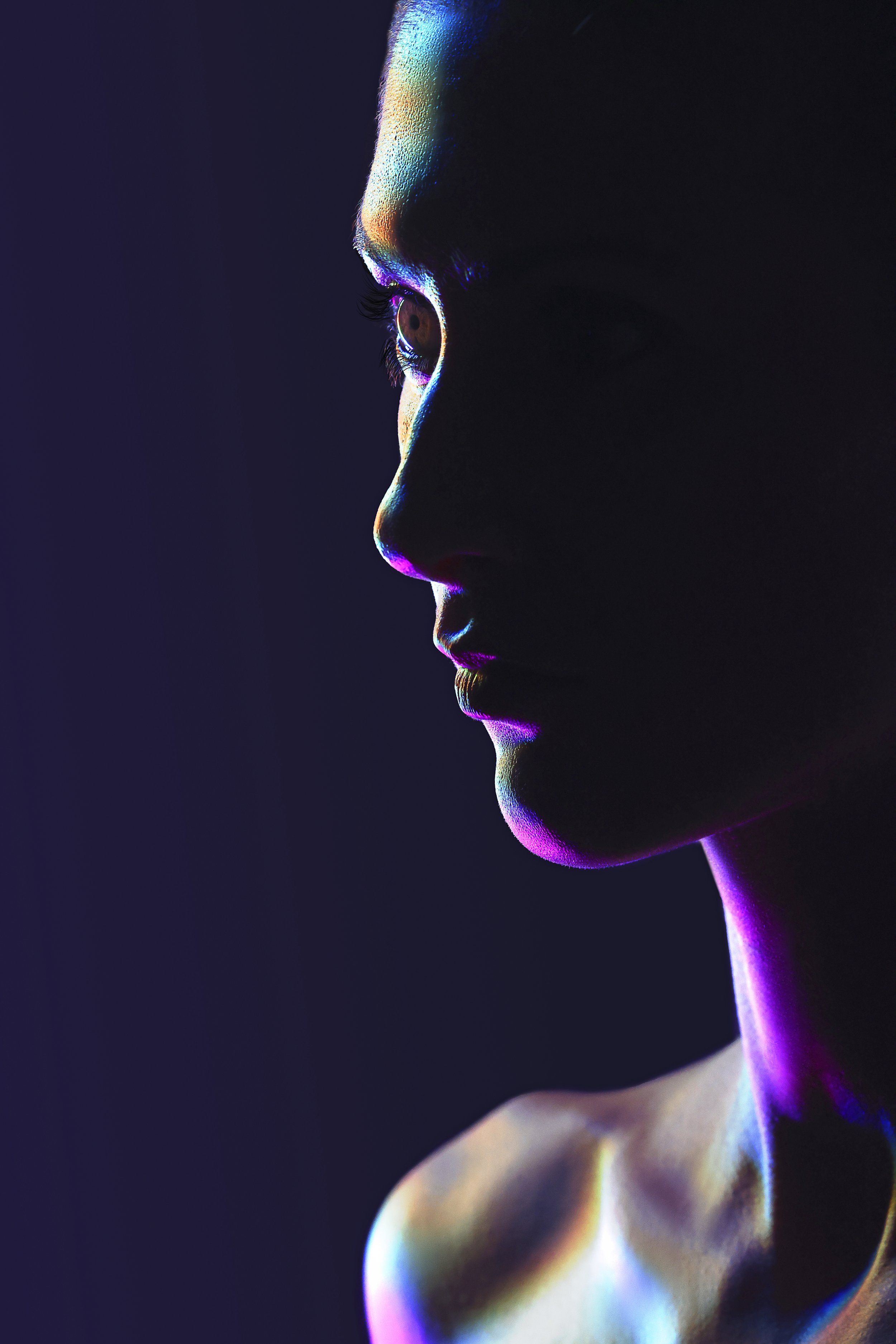 Profile of a woman with colorful neon highlights on her face and shoulder against a dark background.