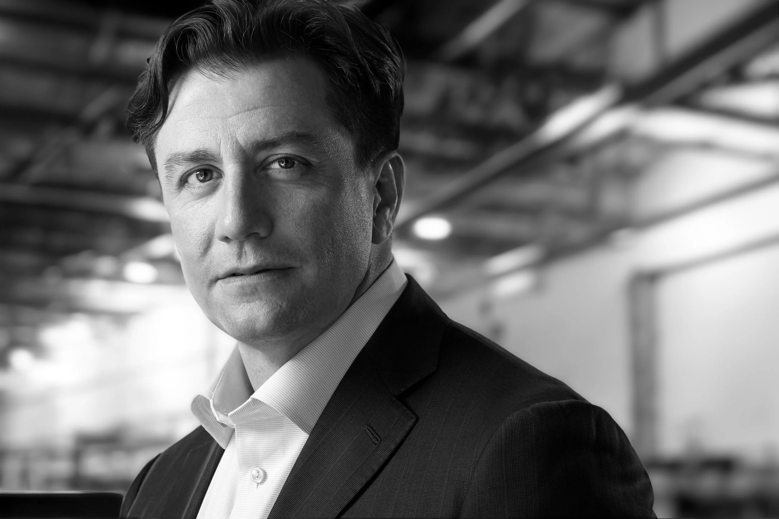 Black and white portrait of a man in a suit, looking directly at the camera with a slight smile, inside a modern office or industrial space.