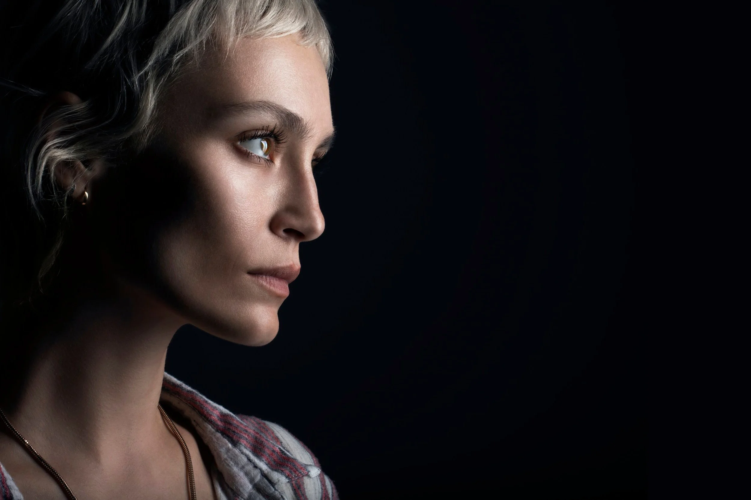 A woman with short, blonde hair, wearing a plaid shirt and a necklace, is shown in profile against a dark background.  Image by top Los Angeles photographer Kenneth Dolin. 