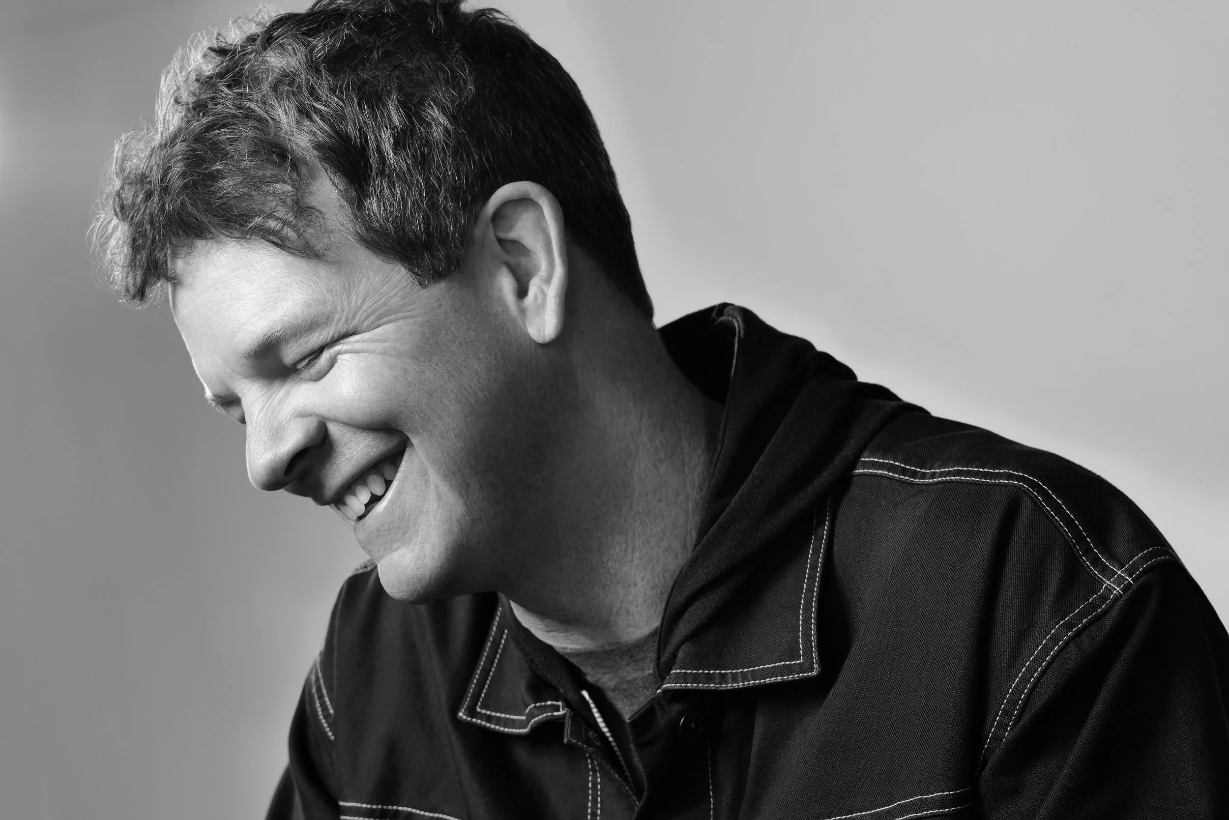 A black and white photo of a man smiling and laughing with his head tilted down, wearing a dark jacket with white stitching.  Image by top Los Angeles photographer Kenneth Dolin. 