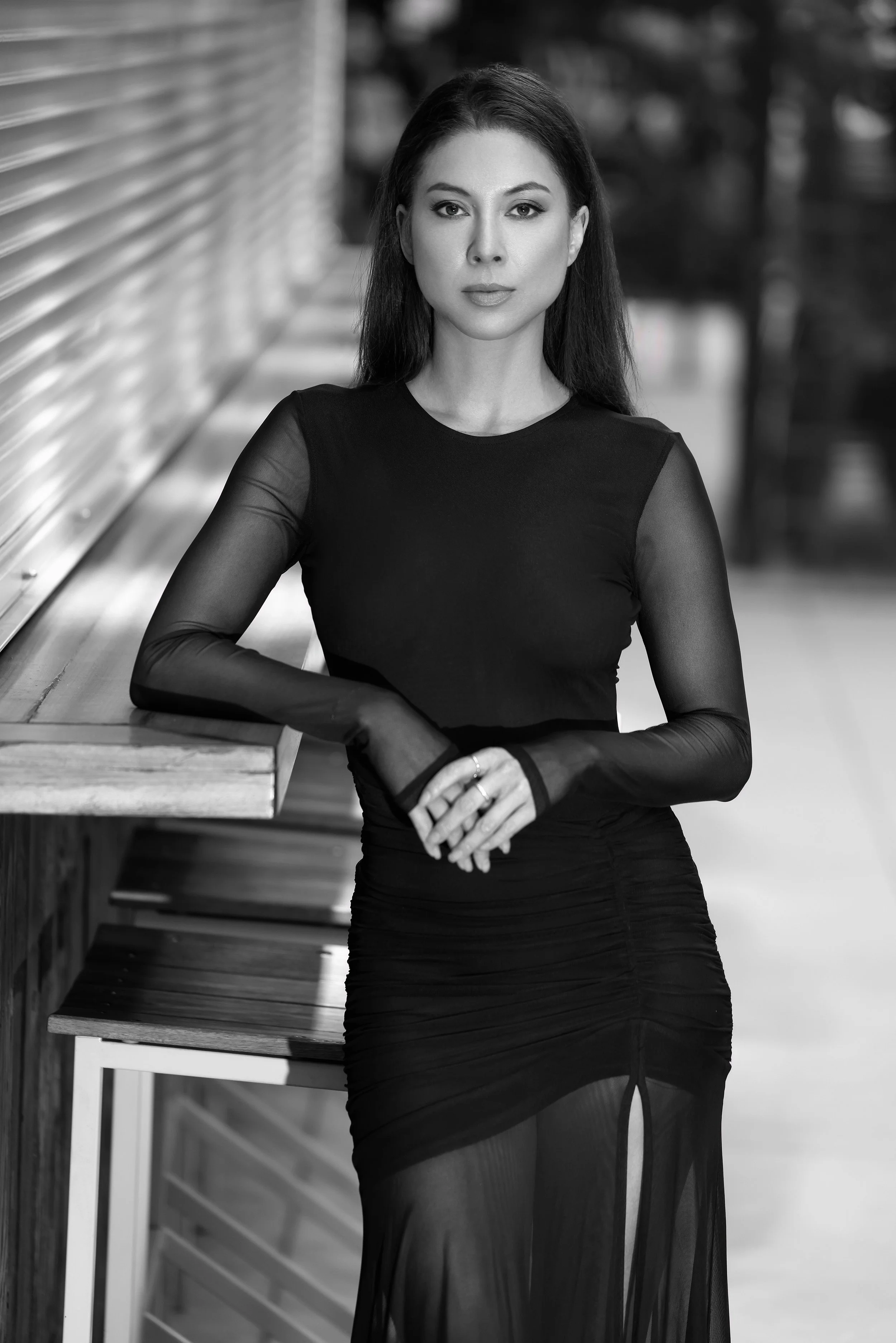 A woman in a black, long-sleeved dress with sheer sleeves, leaning on a wooden counter outdoors.
