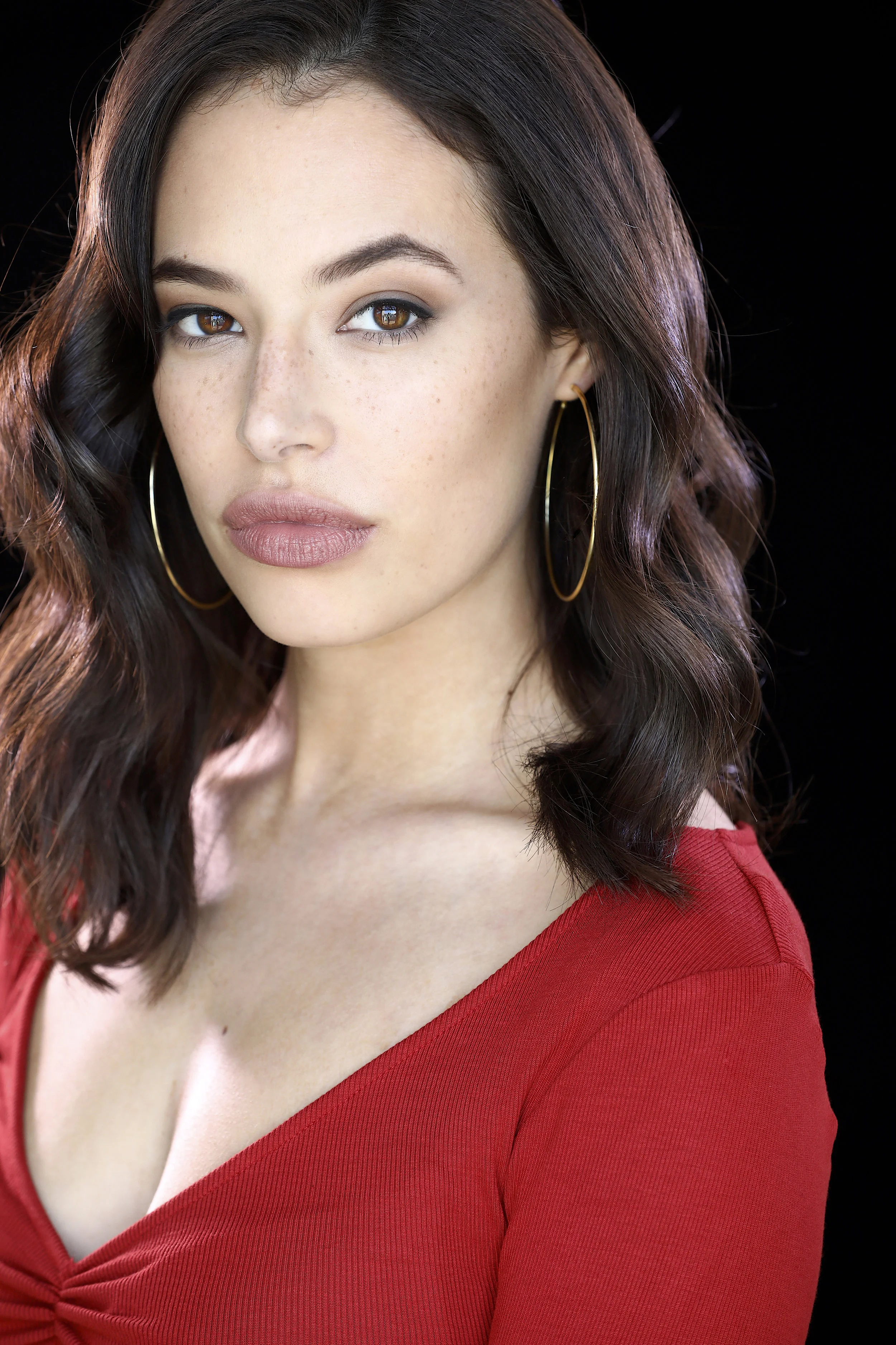 Close-up of a woman with brown eyes and dark wavy hair wearing hoop earrings and a red top against a black background.  Image by top Los Angeles photographer Kenneth Dolin. 