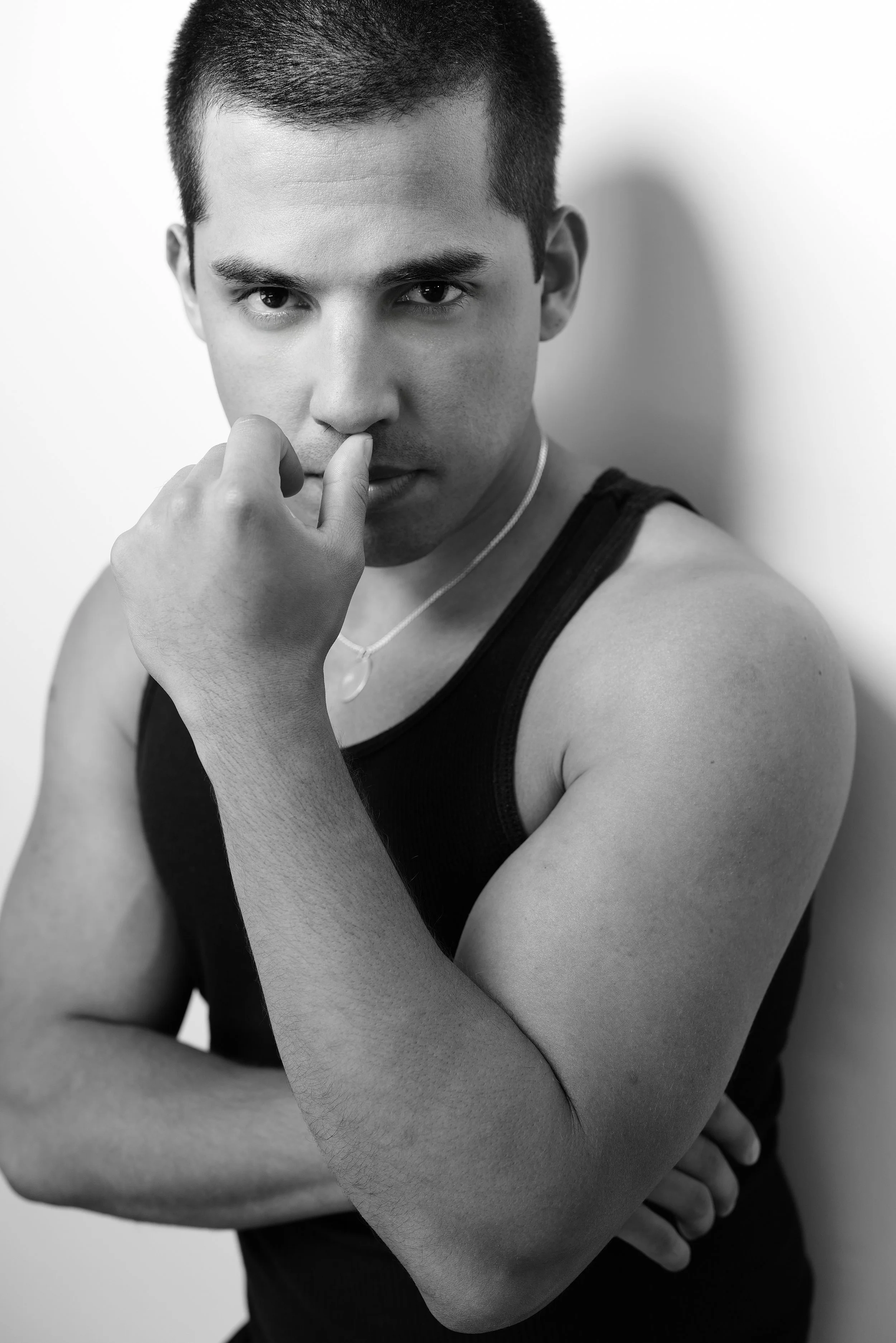 A young man with a serious expression, wearing a black tank top and a necklace, standing against a plain wall, with short dark hair, resting his hand near his face.  Image by top Los Angeles photographer Kenneth Dolin. 