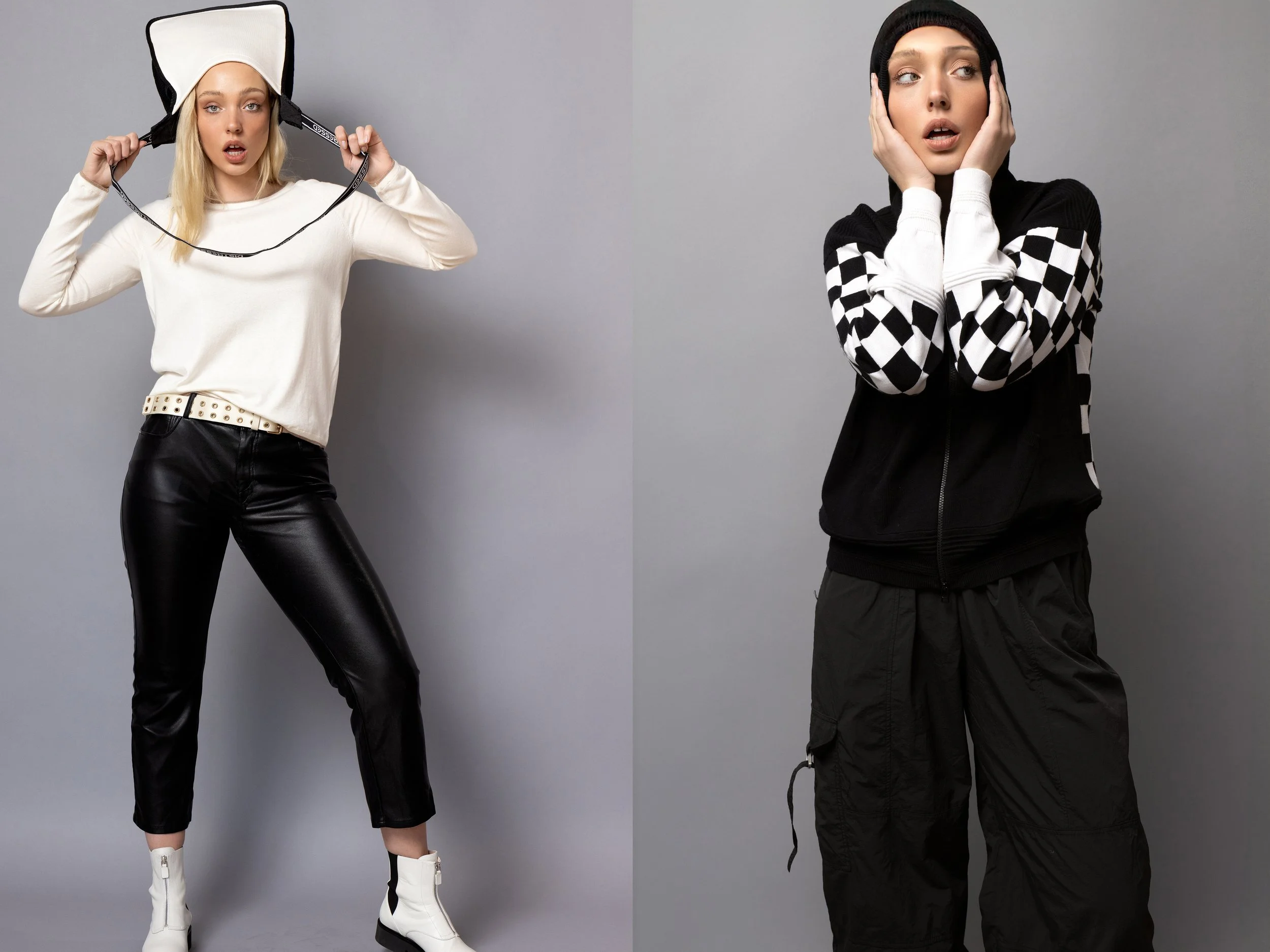 Two women standing against a gray background; the woman on the left is wearing a white sweater, black leather pants, and a black and white helmet, holding the helmet strap with surprised expression; the woman on the right is wearing a black and white