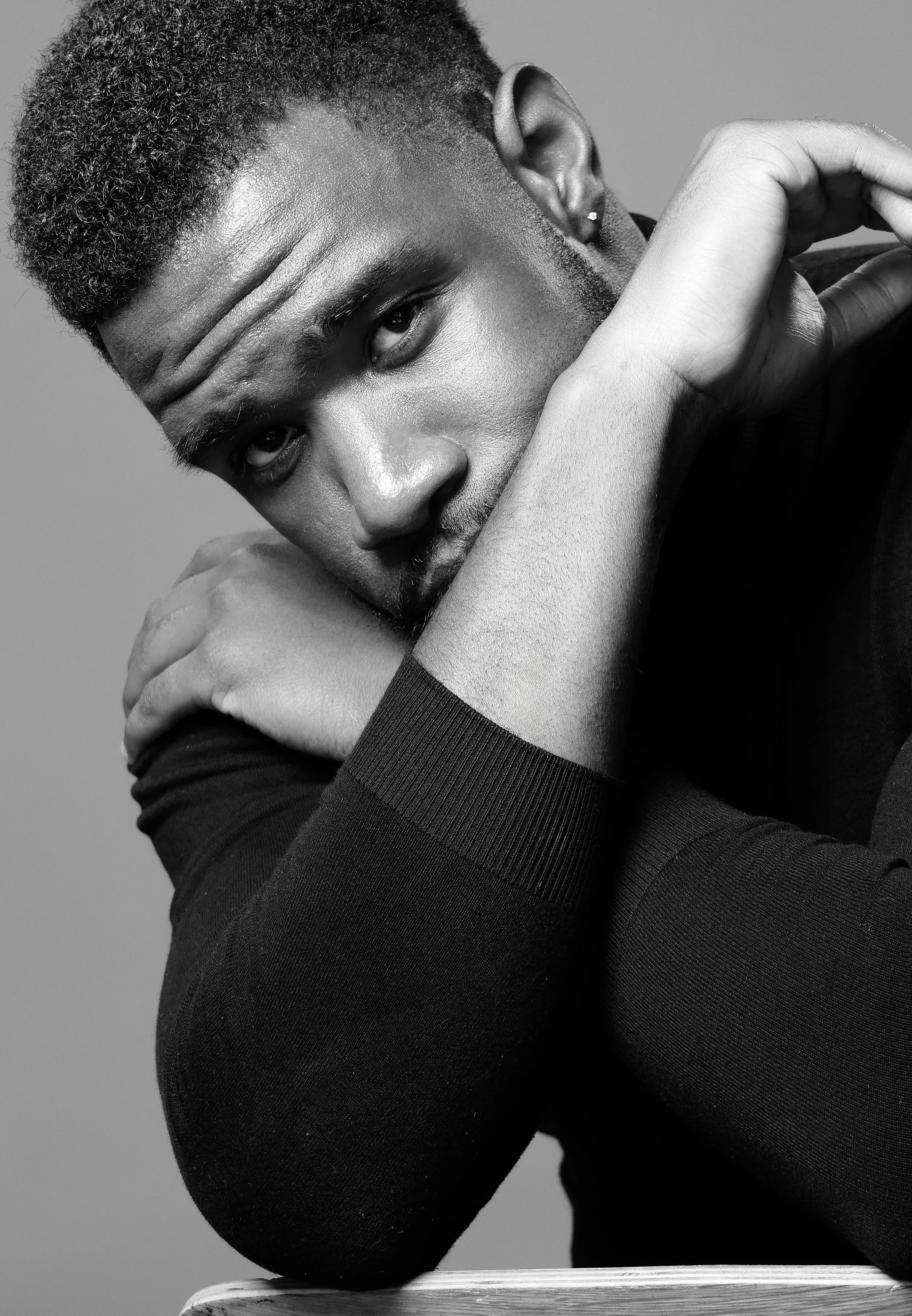 Black and white portrait of a young man with short curly hair resting his head on his crossed arms, looking into the camera.