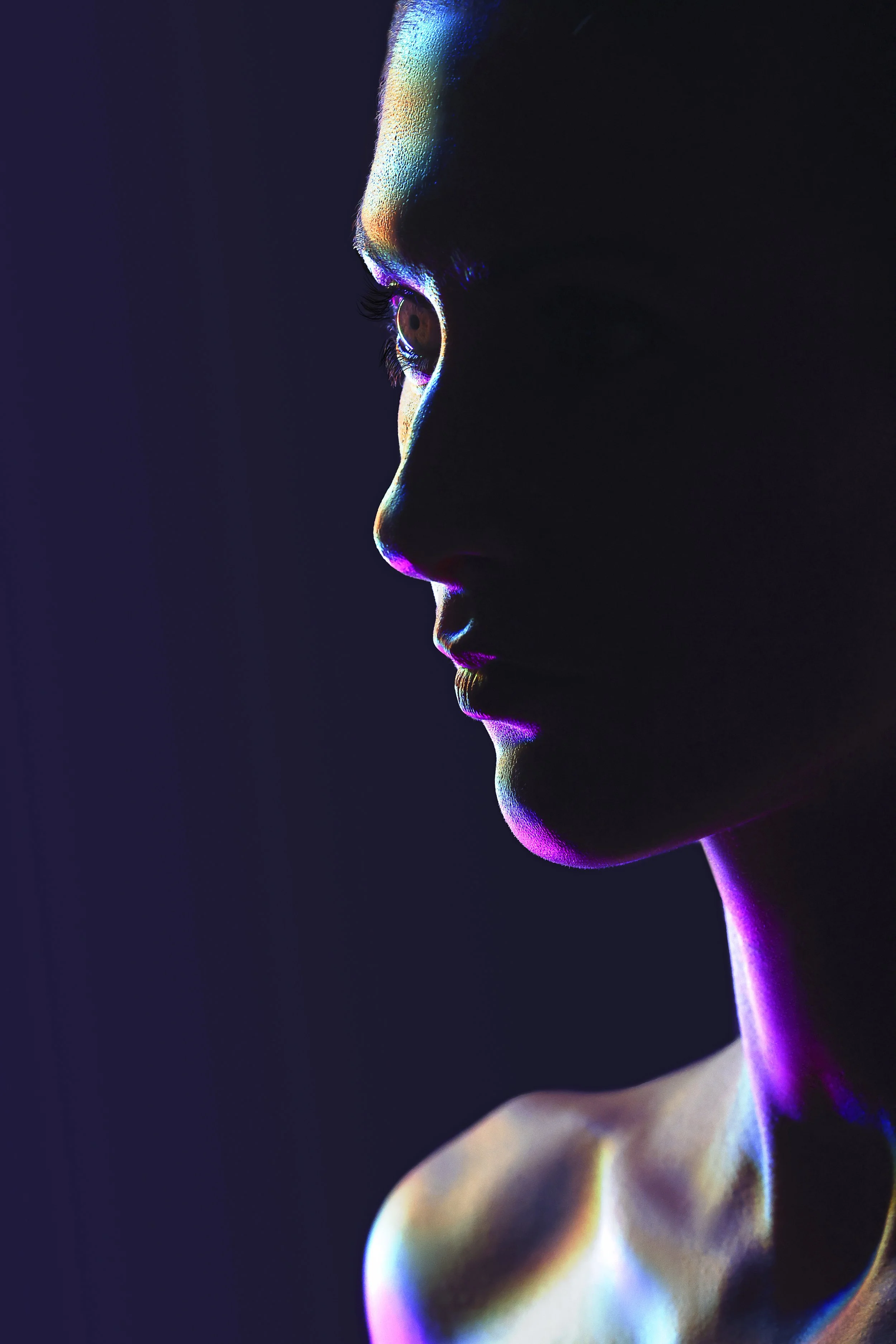Profile of a woman with colorful neon highlights on her face and shoulder against a dark background.