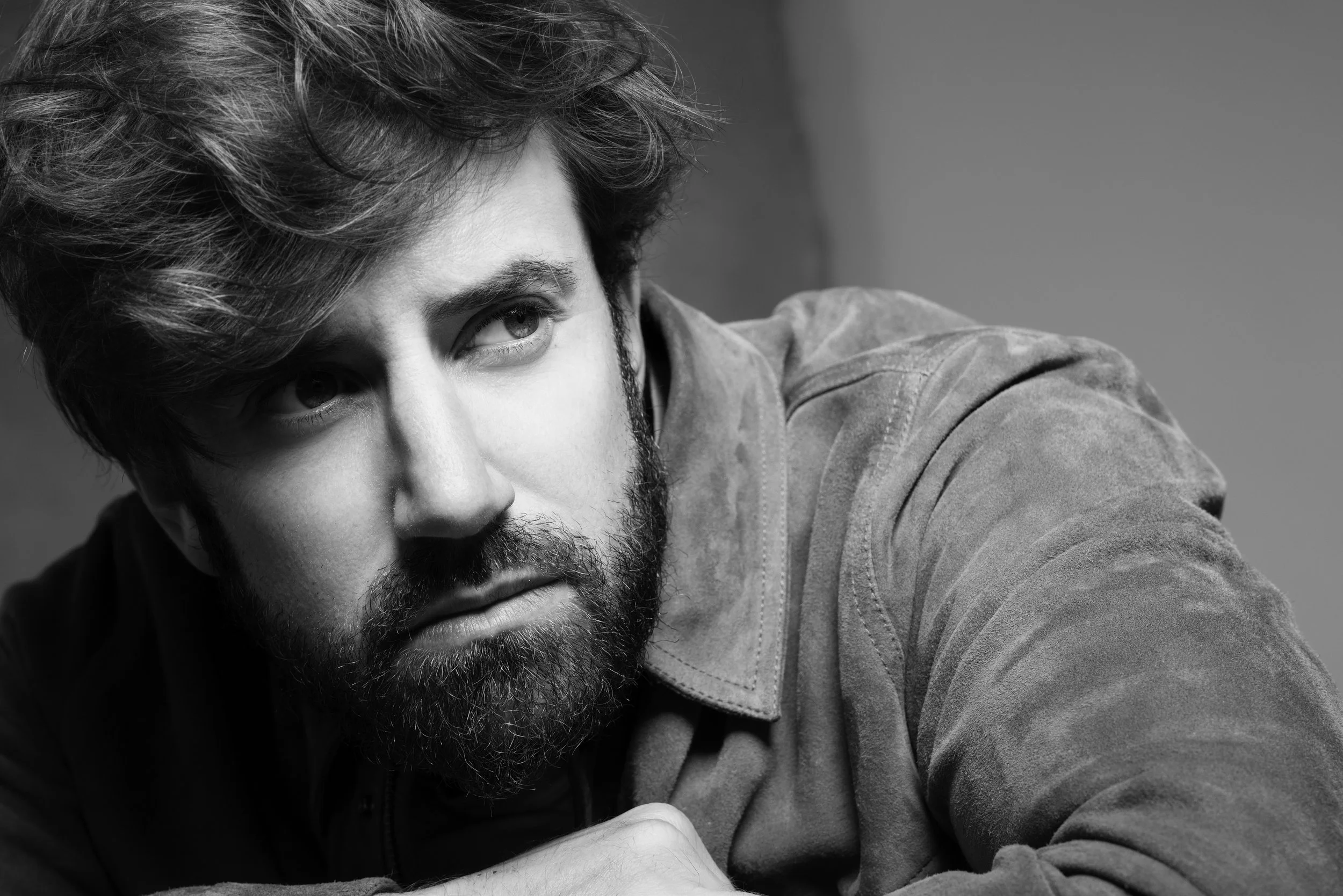 Black and white photo of a young man with curly hair and a beard, resting his head on his arm, looking to the side.